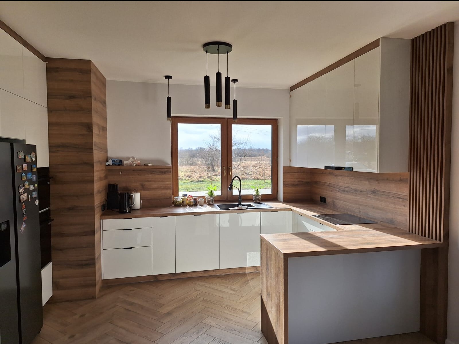 Nowoczesna kuchnia z białymi szafkami i drewnianym blatem. Uwagę przyciąga okno z widokiem na zewnątrz i lampy wiszące nad zlewem. Parkiet w jodełkę.
