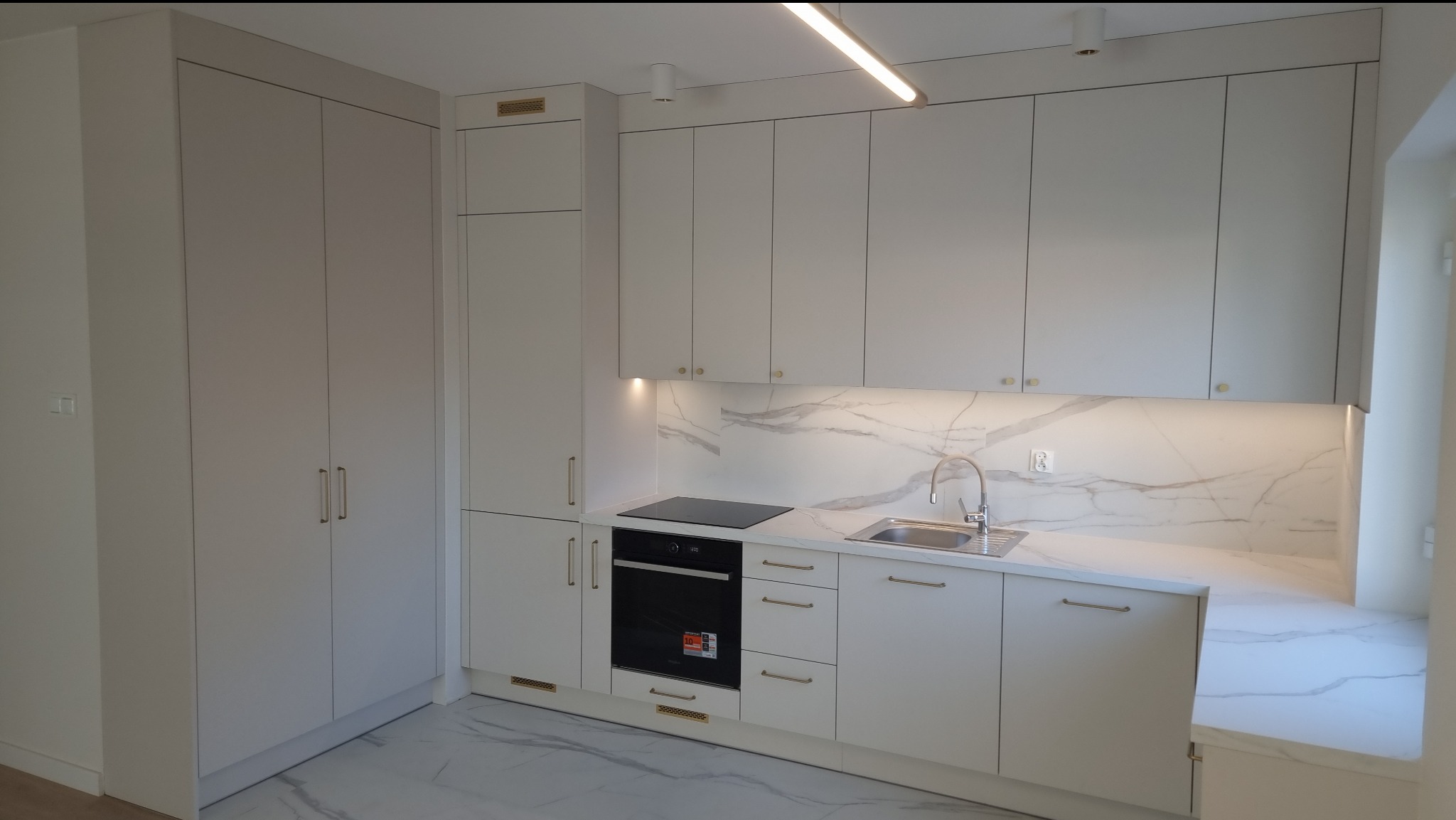 Nowoczesna, jasna kuchnia z zabudową na wymiar: szafki górne i dolne, wysoka szafa, marmurowy backsplash, płyta indukcyjna i piekarnik w zabudowie, złote uchwyty.