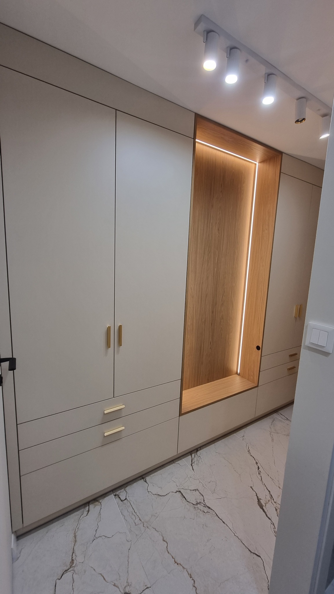 Elegancka szafa wnękowa z beżowymi frontami, złotymi uchwytami i podświetlaną wnęką z drewnianym wykończeniem. Marmurowa podłoga i oświetlenie punktowe.