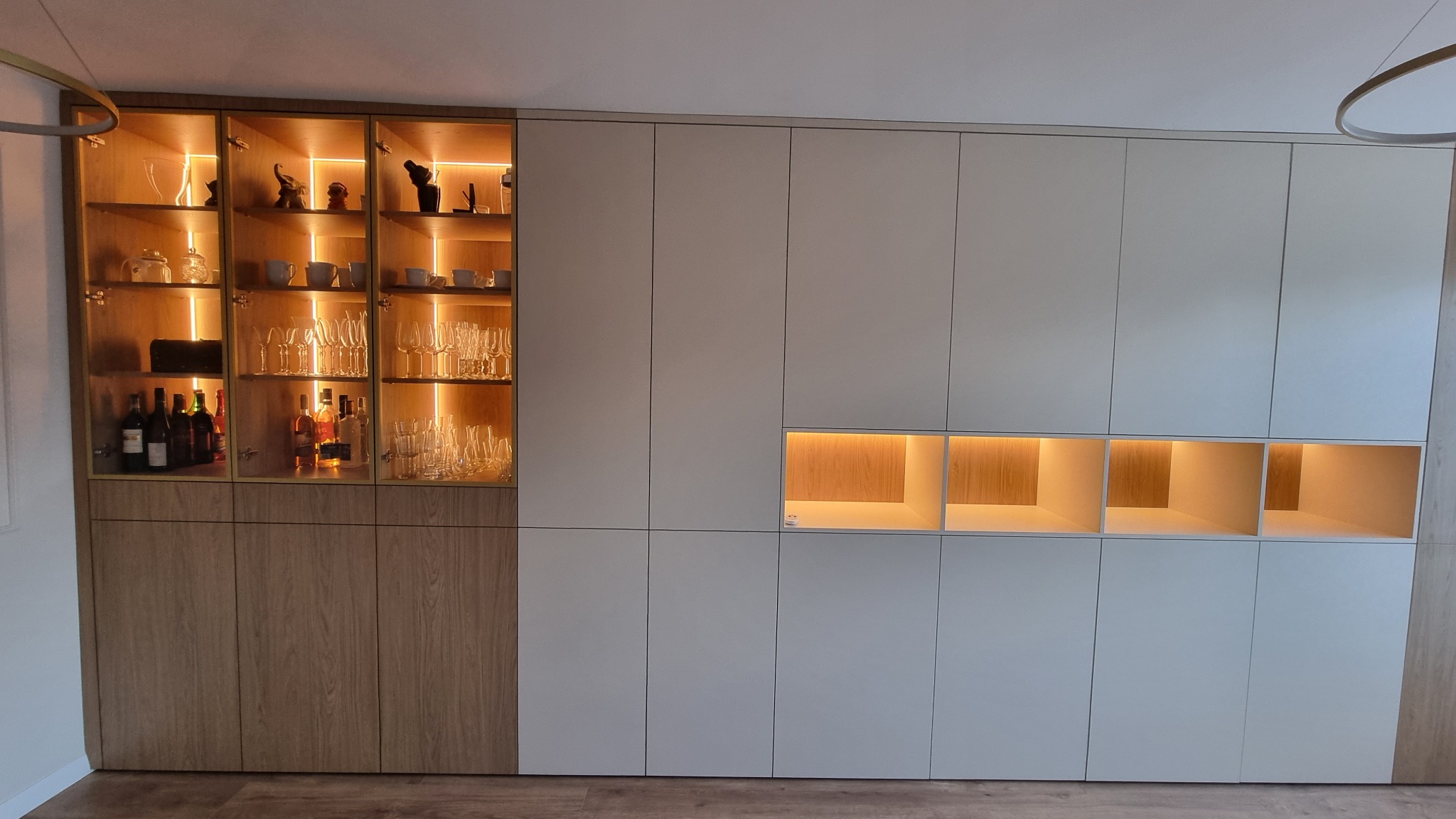 Nowoczesna zabudowa meblowa z podświetlanymi półkami i witryną na alkohole i szkło. Jasne fronty szafek i drewniane akcenty. Oświetlenie LED podkreśla detale.