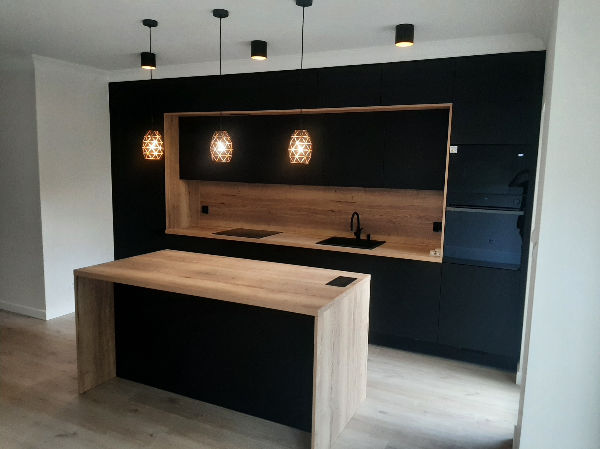 Nowoczesna kuchnia na wymiar z wyspą, czarne fronty i drewniany blat w Gronowie Górnym. Oświetlenie punktowe i wiszące lampy dekoracyjne.