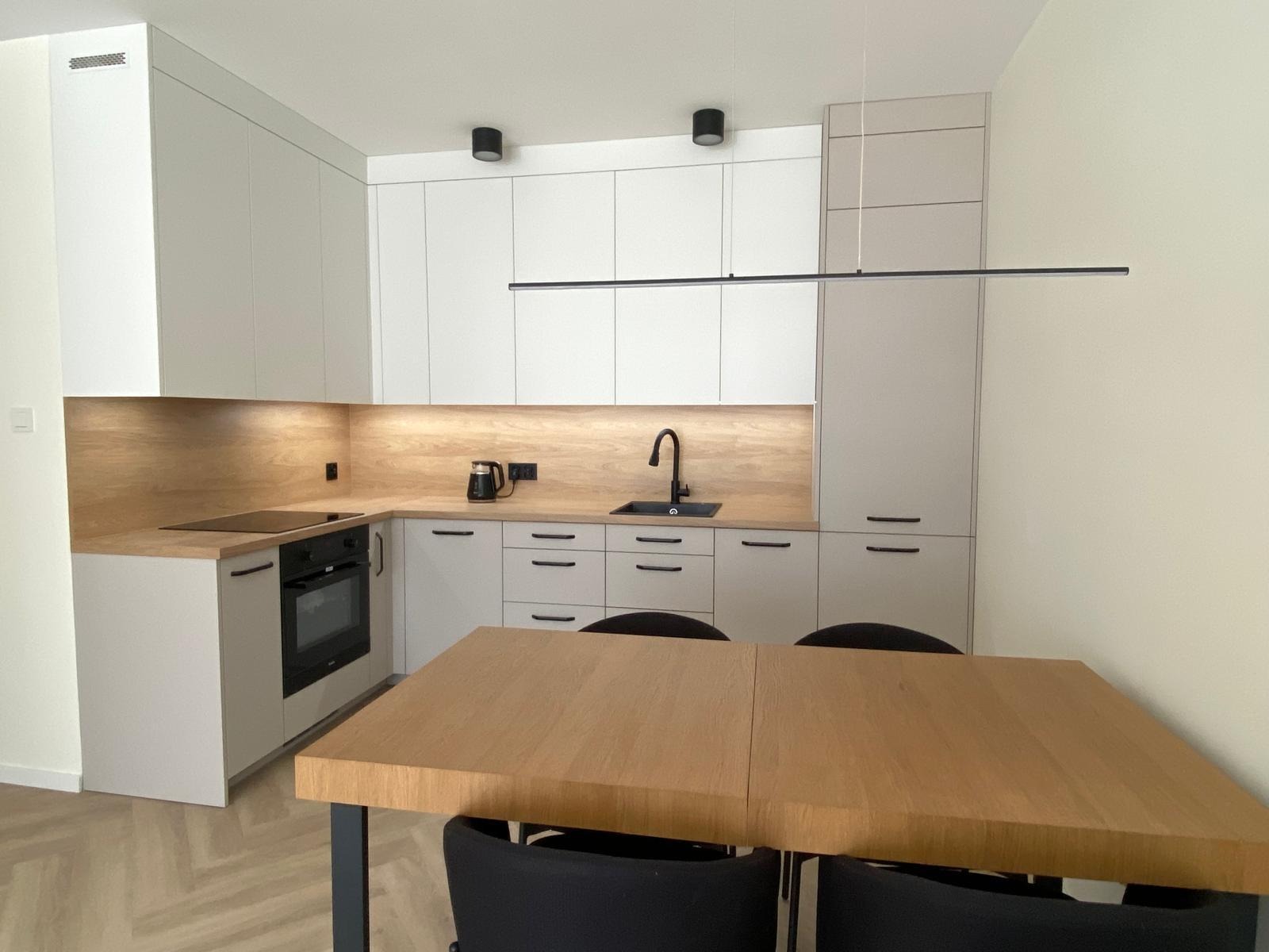 Jasna kuchnia z drewnianym blatem i czarną armaturą, uzupełniona o stół z krzesłami. Nowoczesne oświetlenie punktowe i minimalistyczne uchwyty szafek.