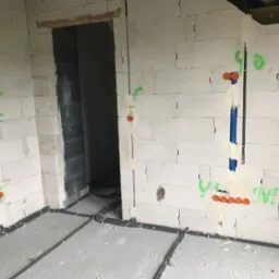 Surowy stan budynku z widocznymi instalacjami elektrycznymi: szare rury prowadzone po podłodze i pionowe przewody w ścianach z betonu komórkowego, oznaczone zielonymi napisami i pomarańczowymi...