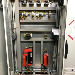 AMPER Usługi Elektryczne Szymon Olechowski - Otwarta szafa sterownicza z widocznymi elementami instalacji elektrycznej, w tym licznymi przewodami, bezpiecznikami, przekaźnikami, modułami sterującymi w kolorze czerwonym oraz dokumentacją...