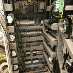 Otwarta szafa sterownicza z licznymi przewodami, przekaźnikami, bezpiecznikami i elementami automatyki, z widocznymi narzędziami i rękawicą roboczą na dolnej półce.