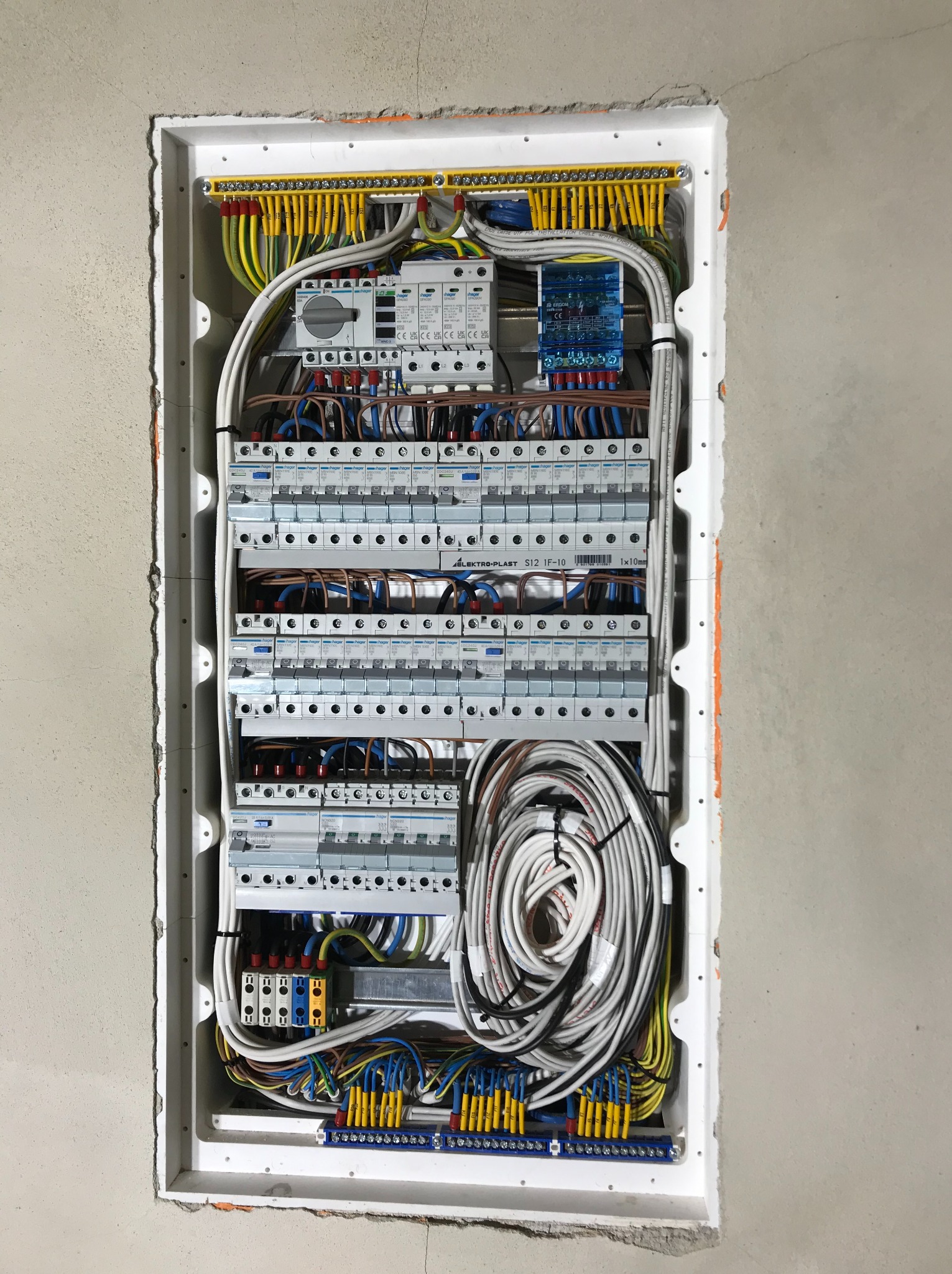 Rozdzielnica elektryczna w białej obudowie, zamontowana w surowej ścianie, widoczne liczne przewody w różnych kolorach, bezpieczniki i inne elementy instalacji.