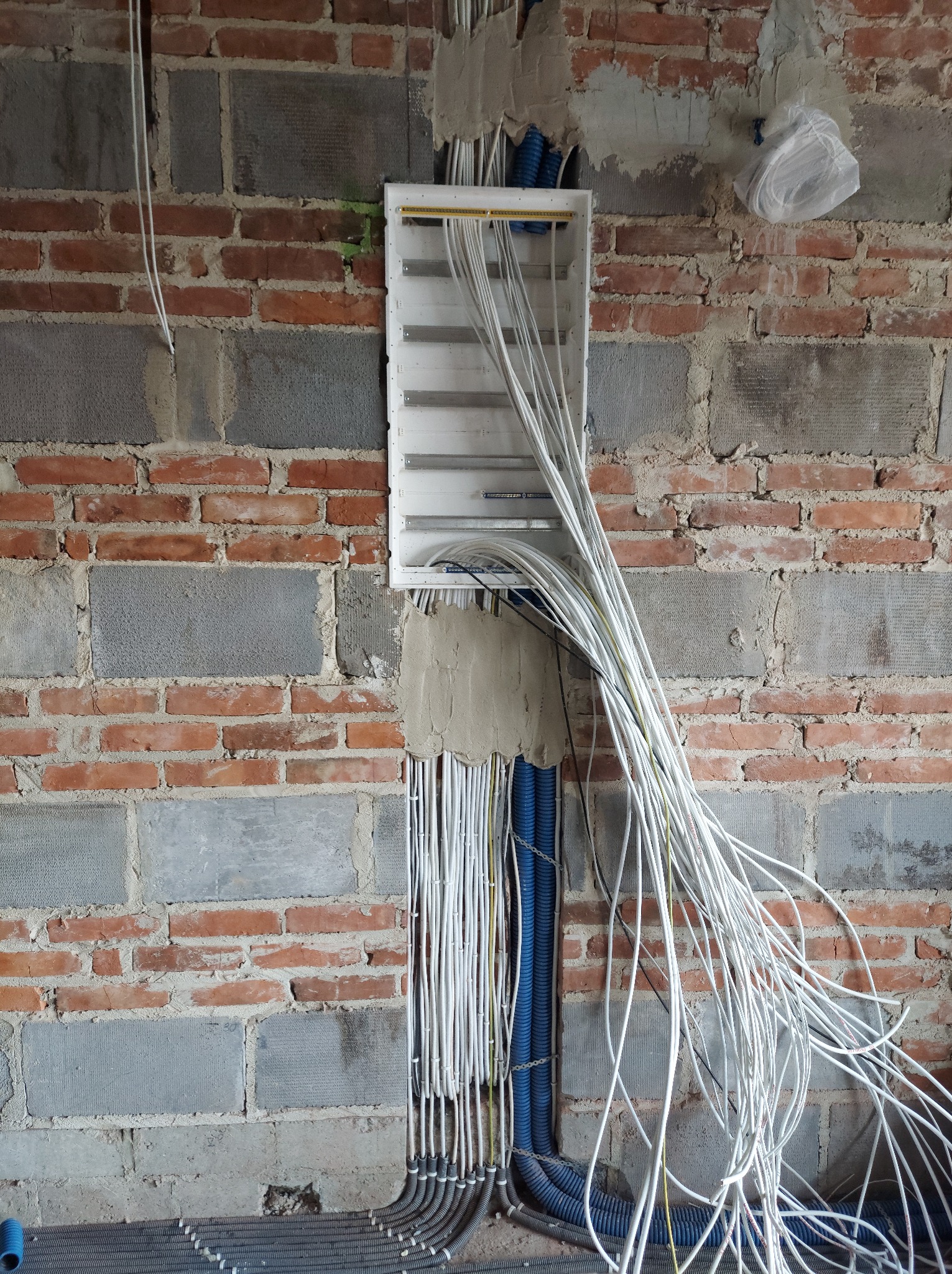 Rozdzielnica elektryczna z wyprowadzonymi przewodami w ścianie z surowej cegły i betonu, widoczne rury karbowane w kolorze niebieskim i szarym.