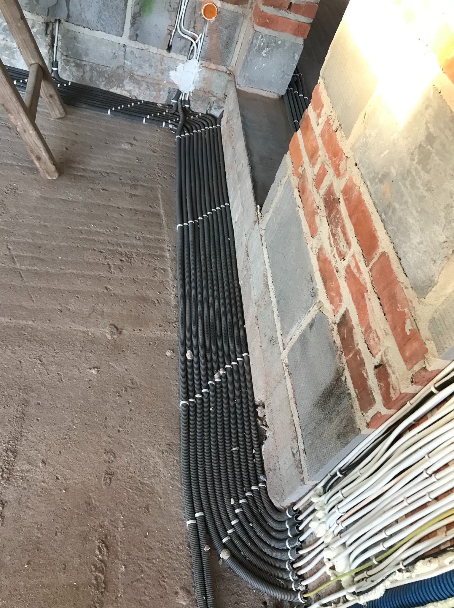 Równolegle ułożone, czarne rury karbowane prowadzące instalację elektryczną wzdłuż surowej ściany z cegły i betonu, z widocznymi białymi przewodami w tle oraz fragmentem drewnianej drabiny.