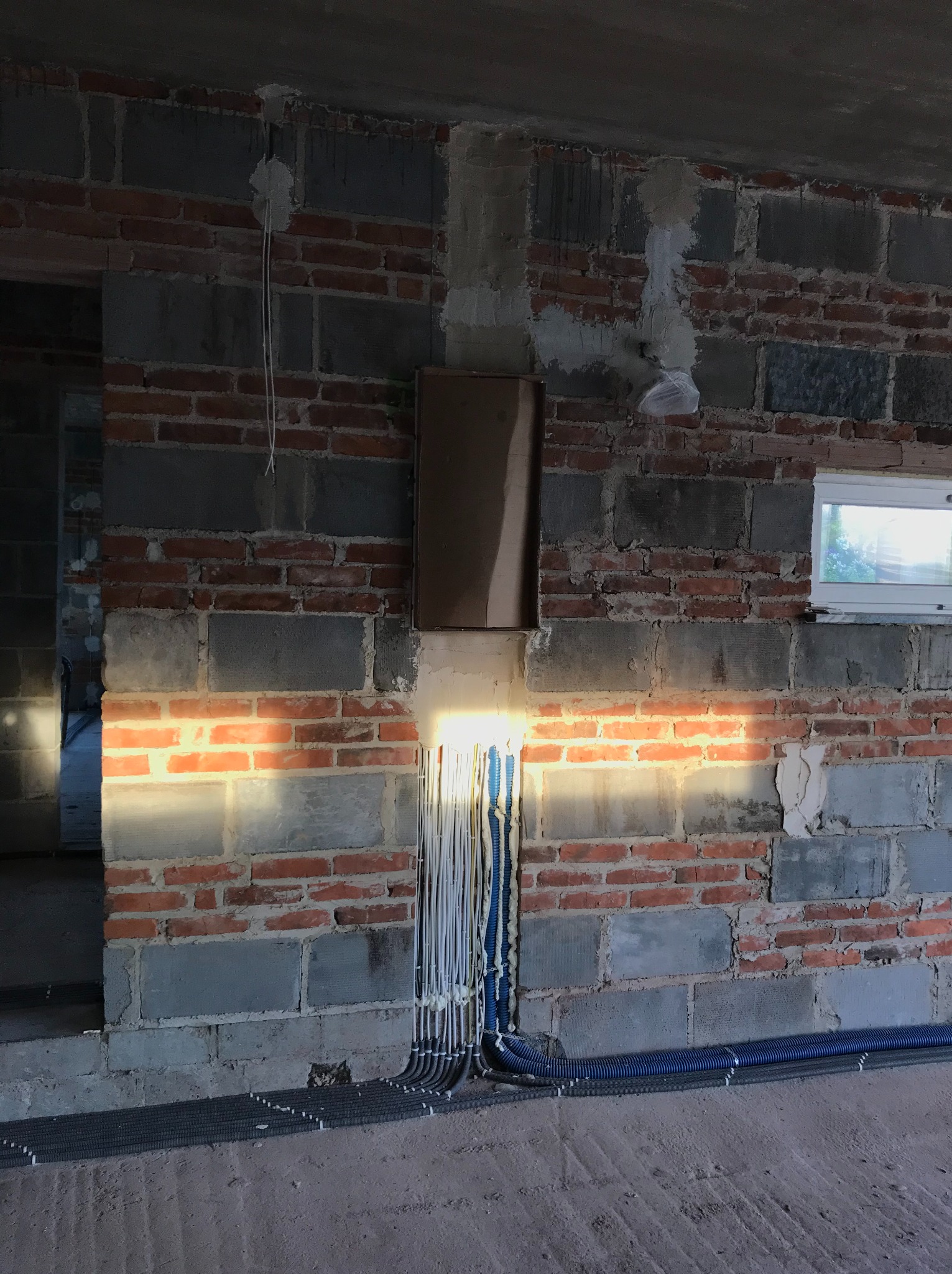 Surowa ściana z cegieł i betonowych bloków w trakcie instalacji elektrycznej, z widocznymi przewodami w peszlach i puszką elektryczną, oświetlona padającym światłem.
