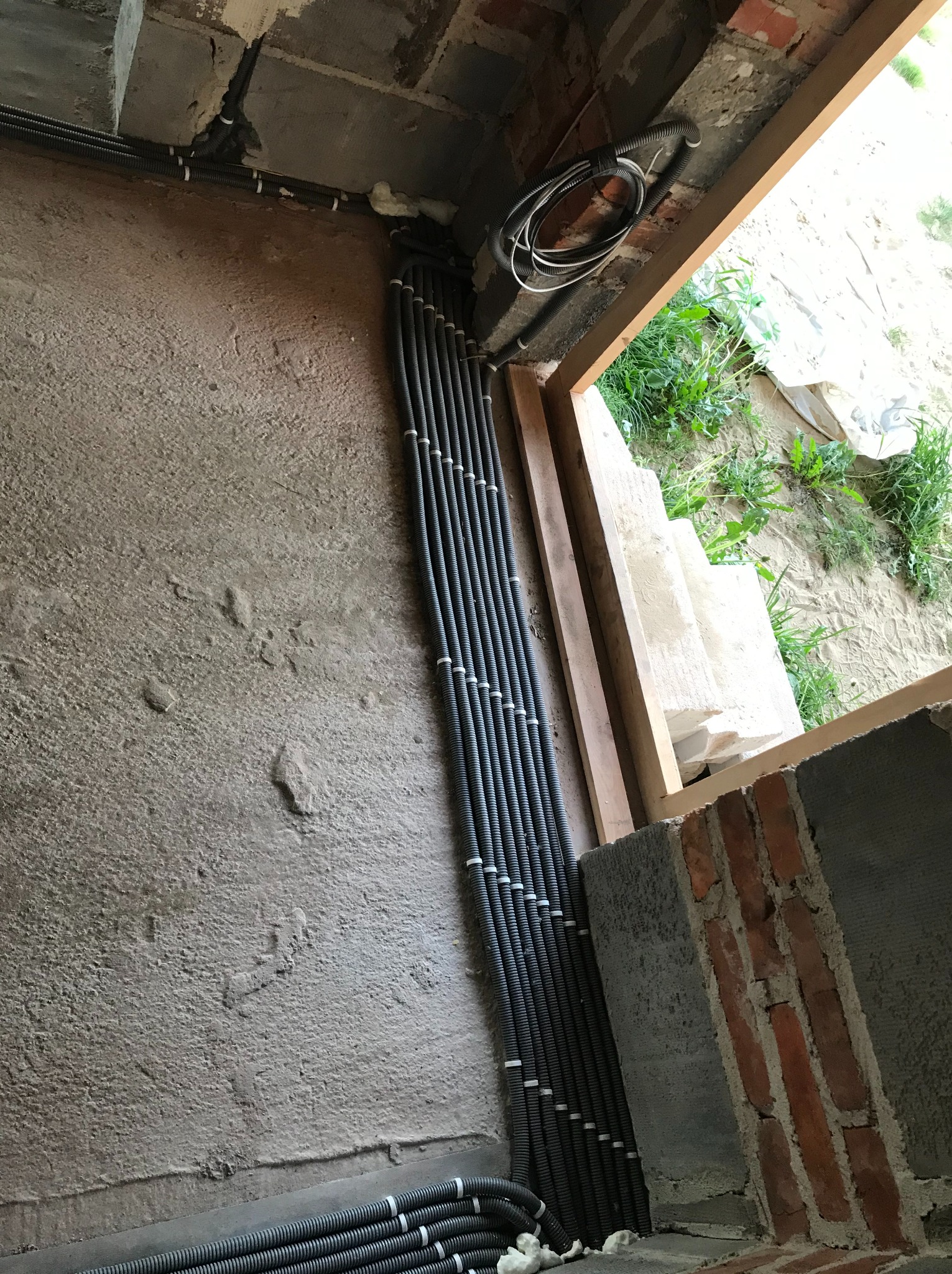 Rzędy czarnych, karbowanych rur instalacyjnych przymocowanych do surowej, betonowej ściany w nowym budynku, z widoczną ramą okienną i fragmentem instalacji na zewnątrz.