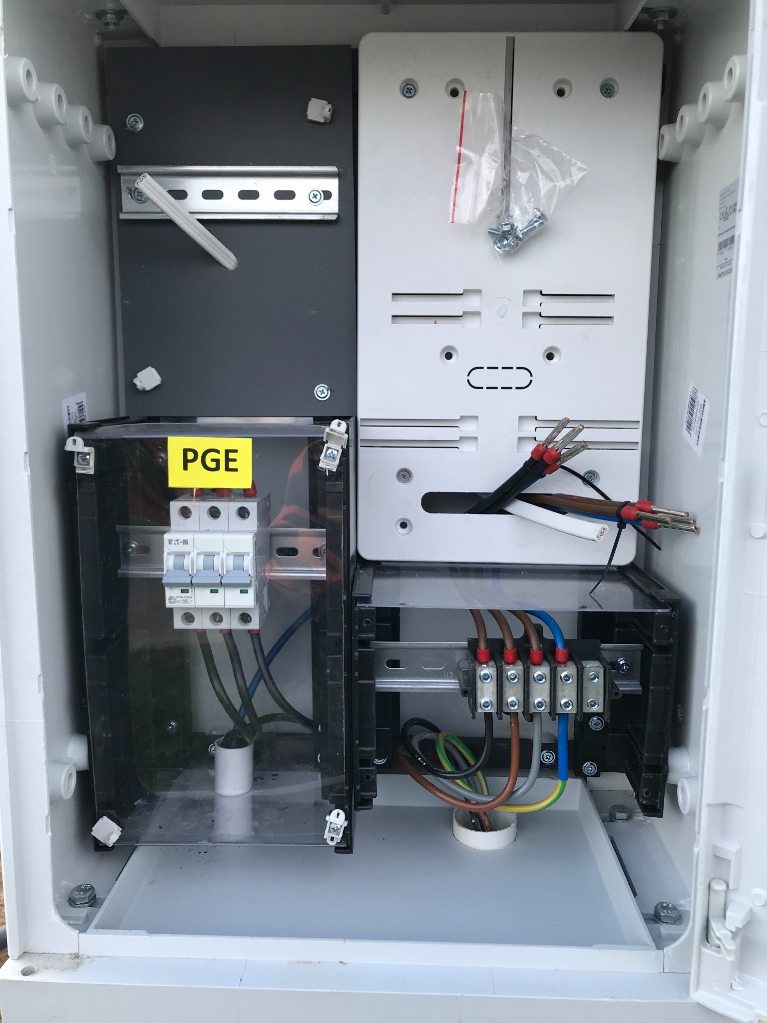 Otwarta skrzynka elektryczna z zabezpieczeniami, przewodami i elementami montażowymi, oznaczona naklejką PGE.