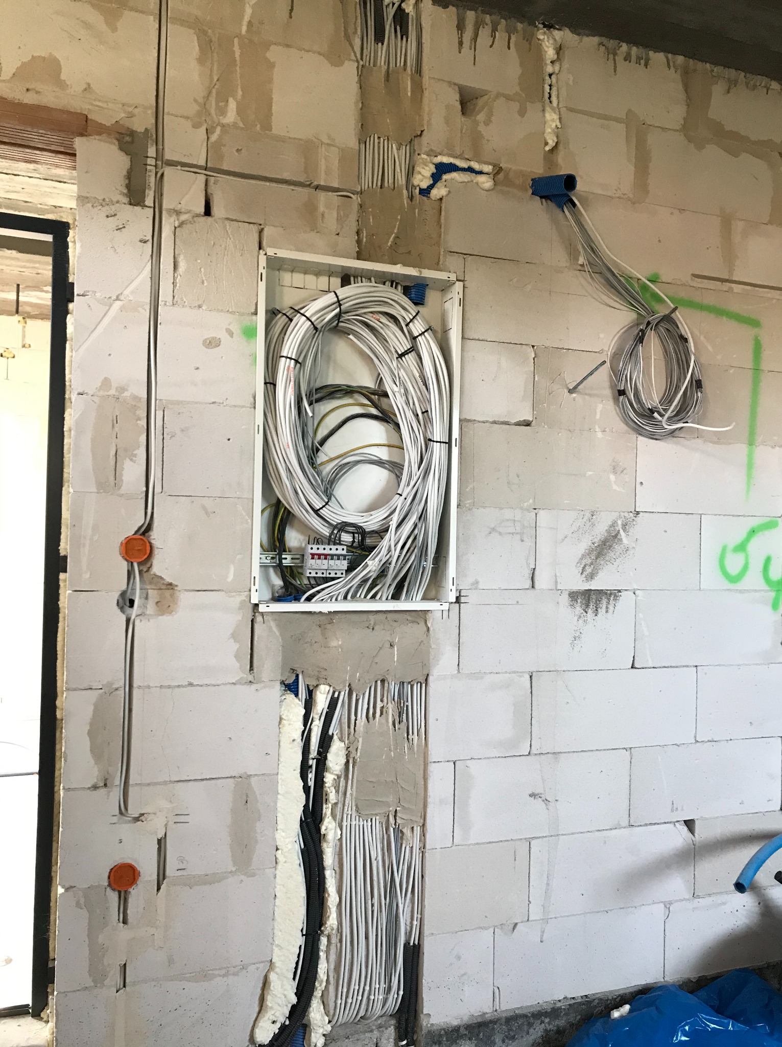 Niedokończona instalacja elektryczna w budynku z białych bloczków, widoczna skrzynka rozdzielcza z licznymi przewodami, peszle z kablami w ścianach, pianka montażowa uszczelniająca przejścia.
