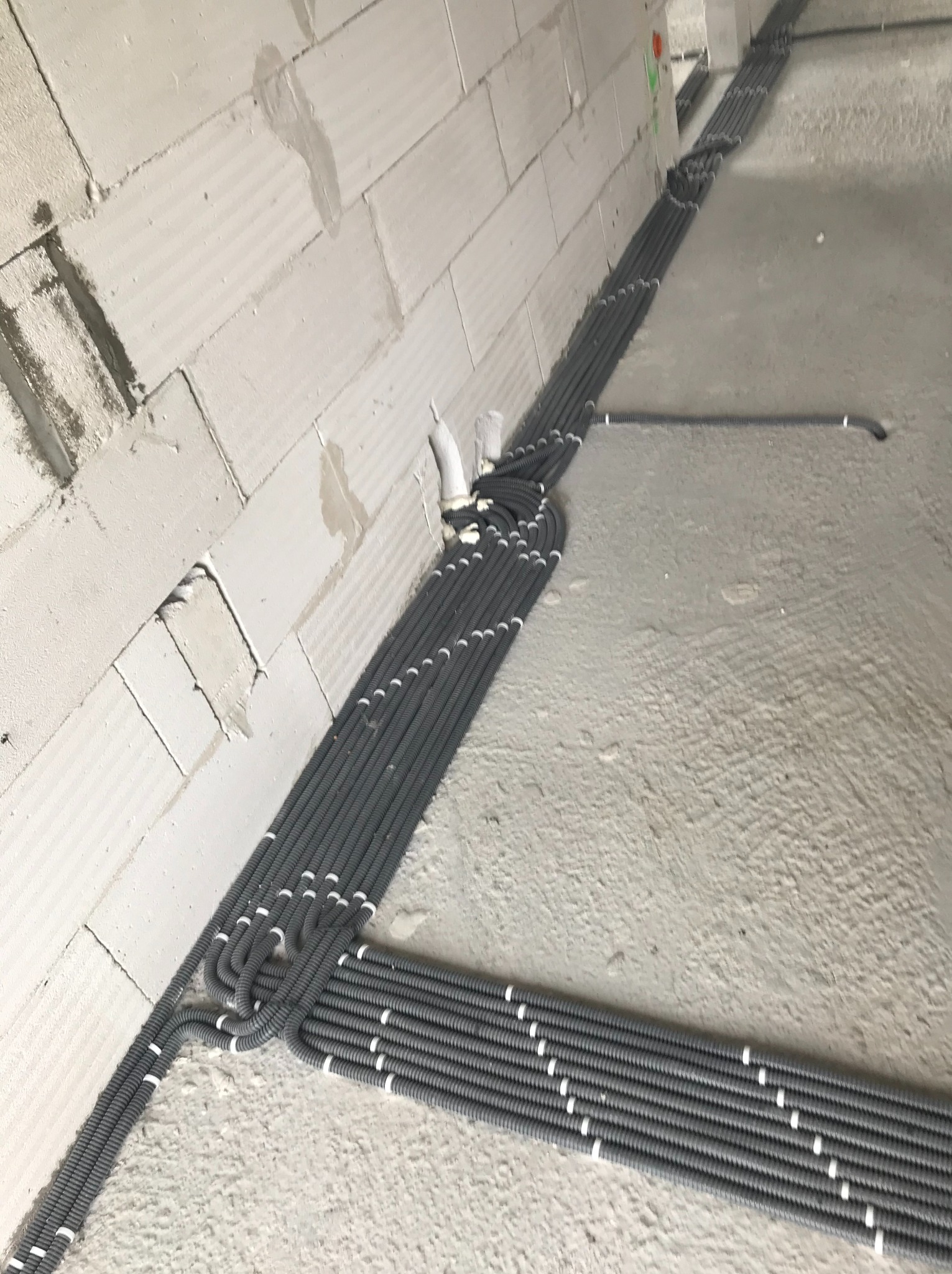 Szare rury karbowane ułożone w wiązki na podłodze i przy ścianie z bloczków w surowym stanie, instalacja elektryczna w trakcie budowy.