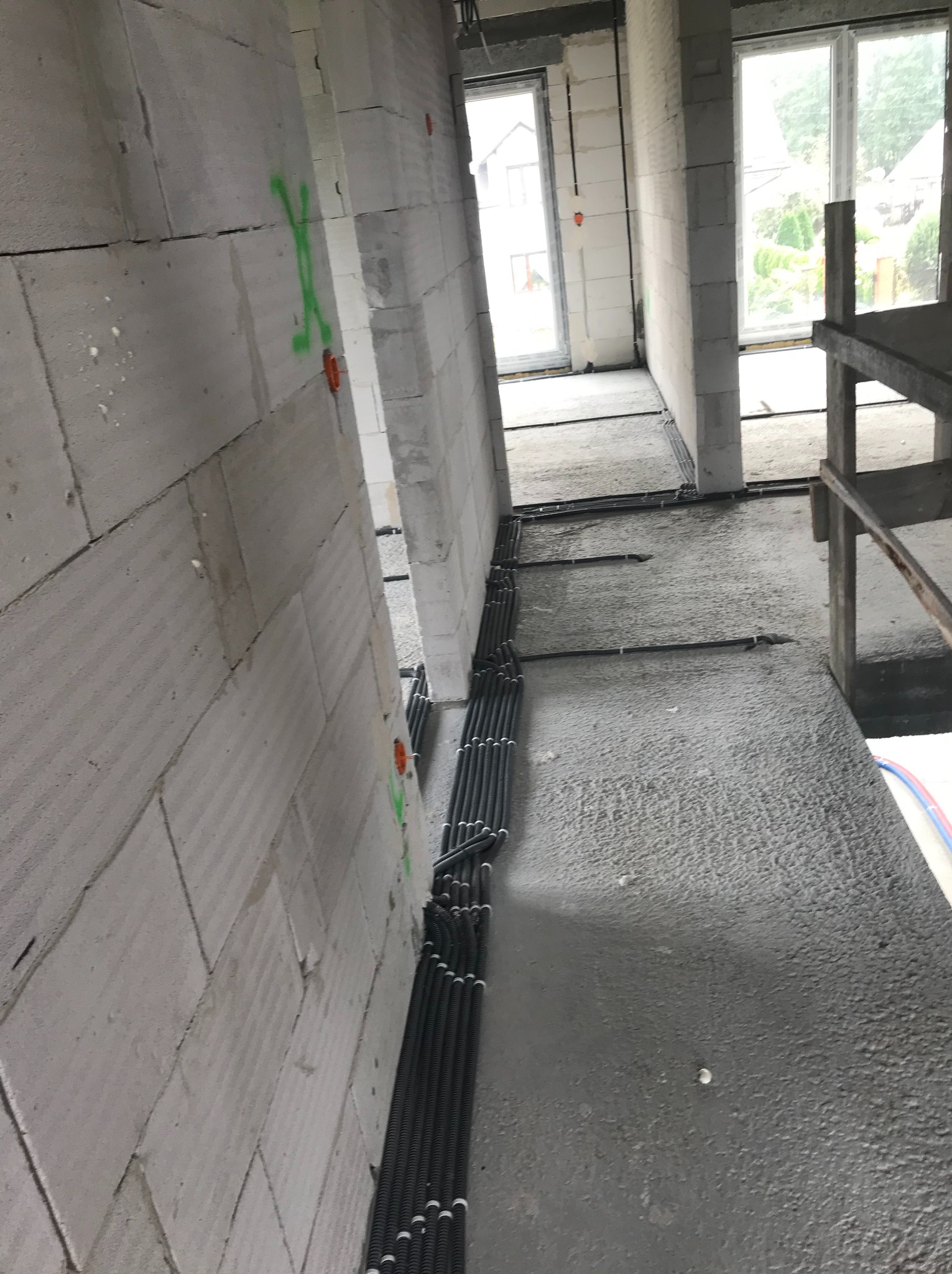 Wnętrze surowego budynku z widocznymi przewodami elektrycznymi w peszlach ułożonymi wzdłuż ściany z betonu komórkowego, z zielonymi oznaczeniami na ścianie.