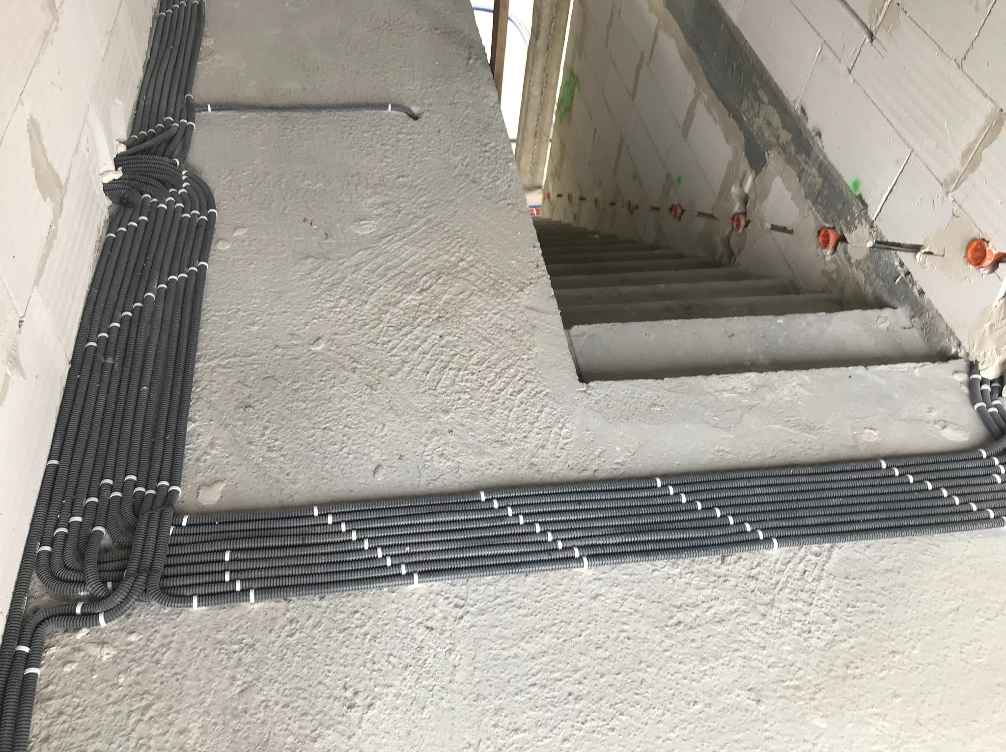 Szare, karbowane rury ochronne na kable elektryczne, ułożone równolegle wzdłuż betonowej ściany i schodów w surowym stanie, z widocznymi puszkami instalacyjnymi w ścianie.
