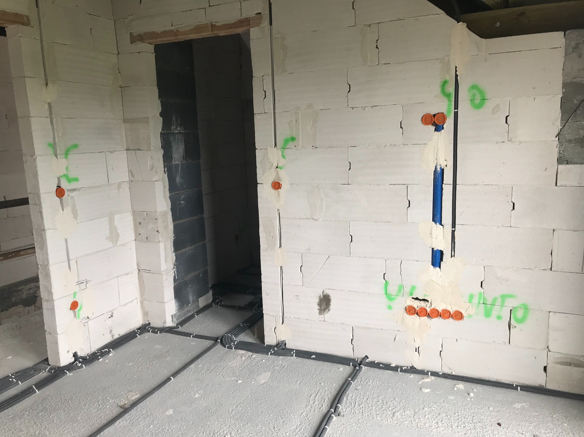 Surowy stan budynku z widocznymi instalacjami elektrycznymi: szare rury prowadzone po podłodze i pionowe przewody w ścianach z betonu komórkowego, oznaczone zielonymi napisami i pomarańczowymi...