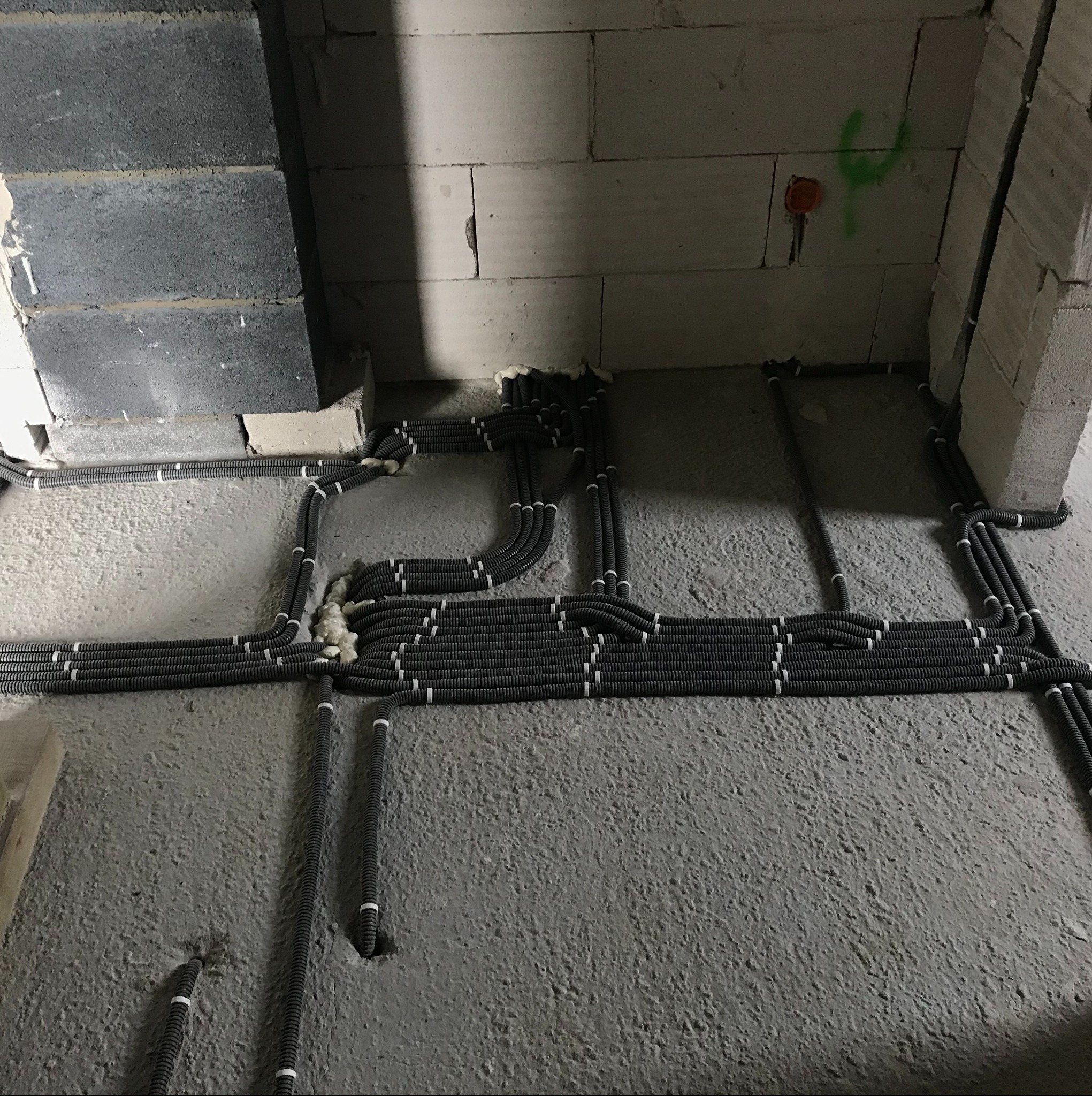 Szare rury karbowane ułożone na surowym betonowym podłożu w trakcie układania instalacji elektrycznej w budynku z szarego betonu komórkowego, widoczne puszki instalacyjne i zielone oznaczenia...
