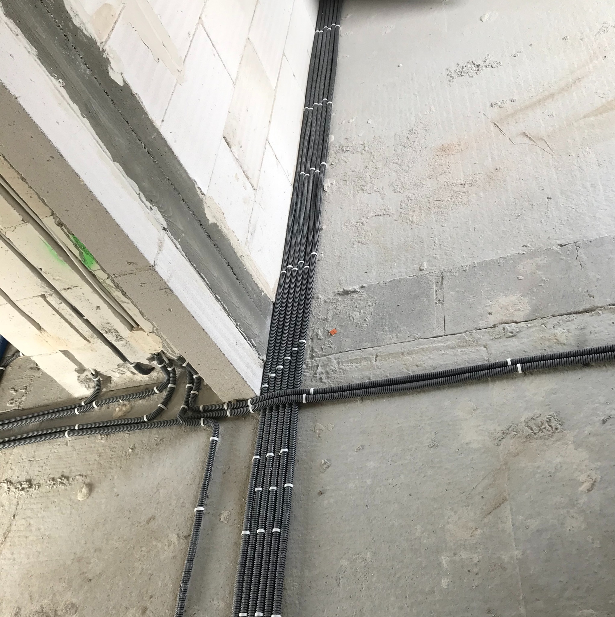 Szereg szarych rurek karbowanych z białymi uchwytami, ułożonych pionowo i poziomo na surowej, betonowej ścianie w trakcie budowy.