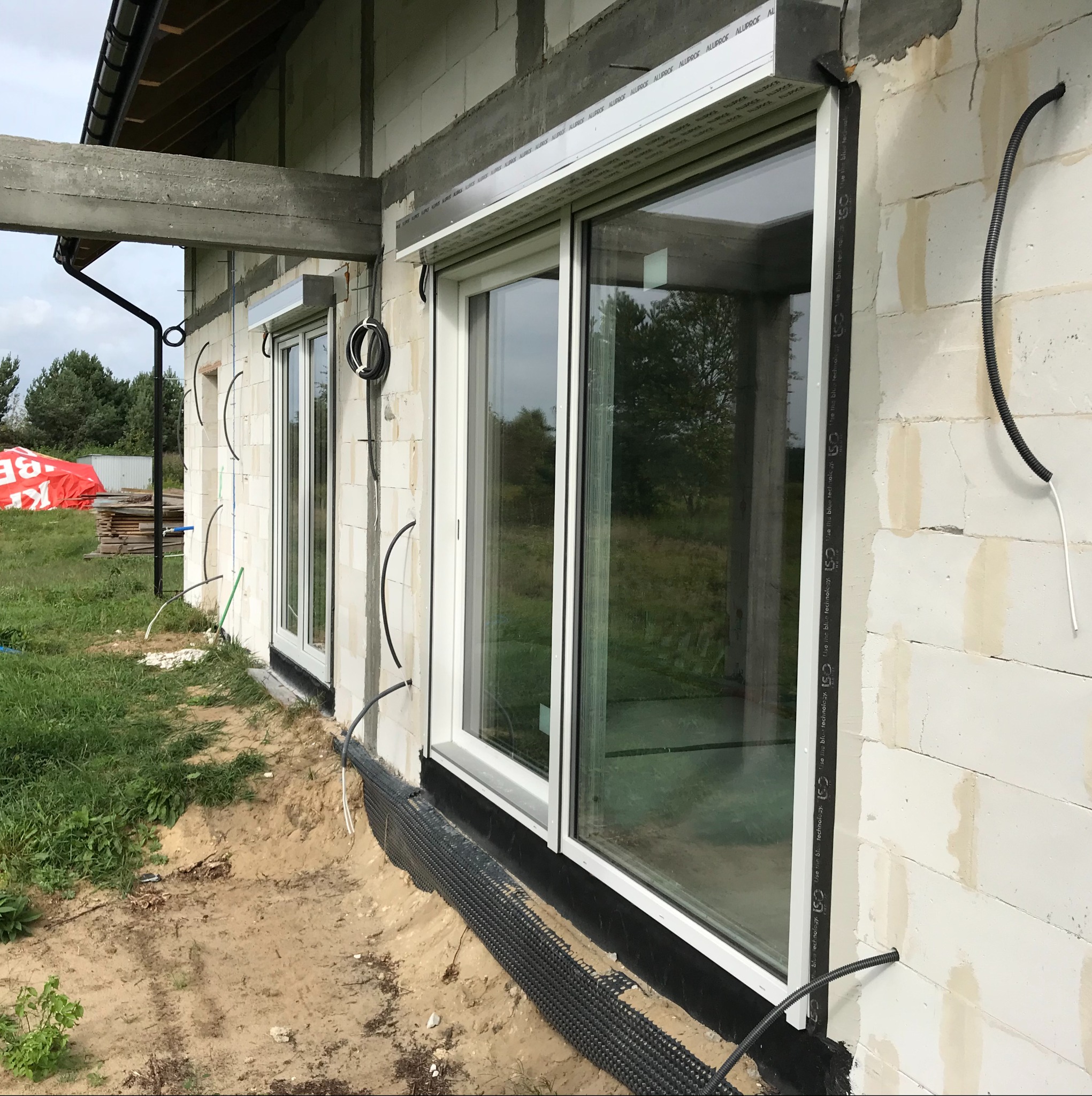 Surowy stan elewacji budynku z oknami i zamontowanymi roletami zewnętrznymi, widoczne peszle elektryczne wystające ze ściany, czekające na podłączenie.