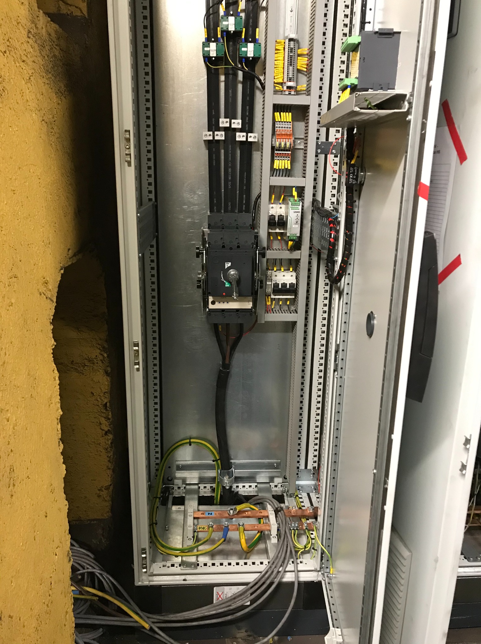 Otwarta szafa elektryczna z widocznymi przewodami, bezpiecznikami i elementami sterującymi, z żółtą ścianą w tle.