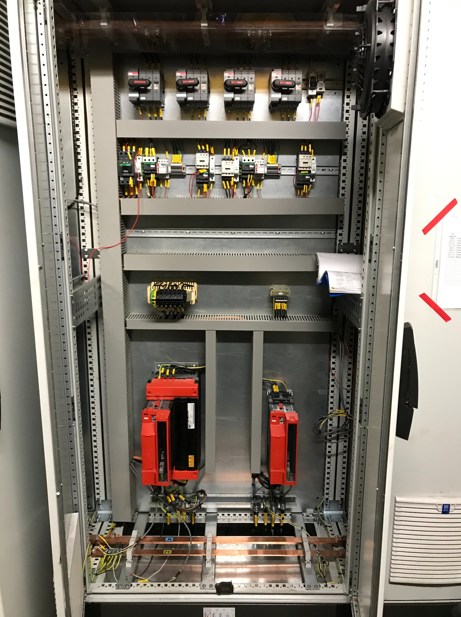 Otwarta szafa sterownicza z widocznymi elementami instalacji elektrycznej, w tym licznymi przewodami, bezpiecznikami, przekaźnikami, modułami sterującymi w kolorze czerwonym oraz dokumentacją...