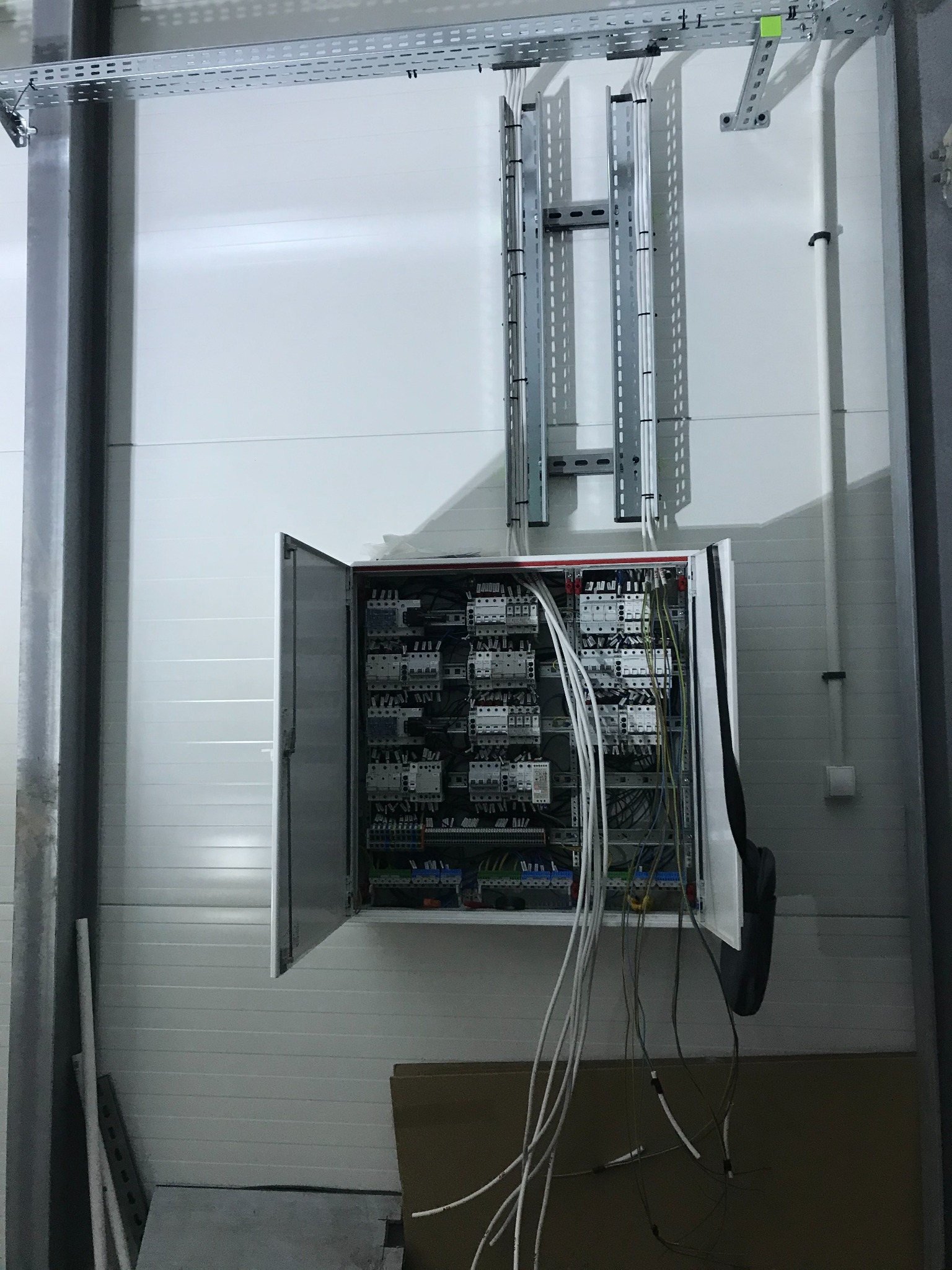 Otwarta szafa sterownicza z widocznymi przekaźnikami, bezpiecznikami i przewodami, zamontowana na białej ścianie z poziomymi panelami, metalowe korytka kablowe nad szafą.