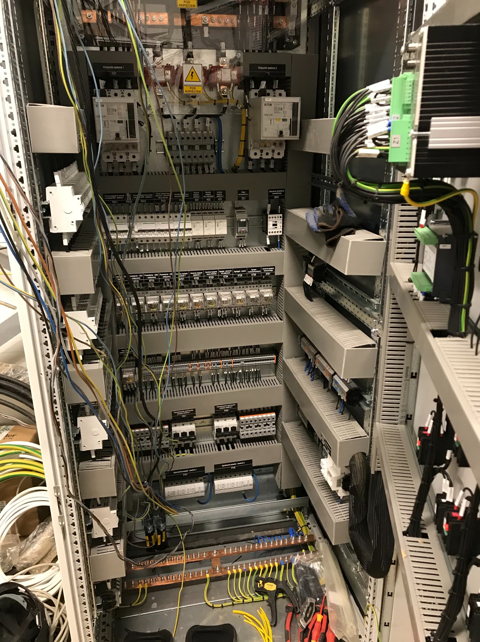 Otwarta szafa sterownicza z licznymi przewodami, przekaźnikami, bezpiecznikami i elementami automatyki, z widocznymi narzędziami i rękawicą roboczą na dolnej półce.