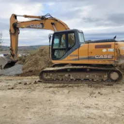 Pomarańczowa koparka CASE CX210B na placu budowy, z łyżką wypełnioną drobnym kruszywem, widoczne ślady błota na gąsienicach i dolnej części maszyny, w tle hałda ziemi i zalesione wzgórza...