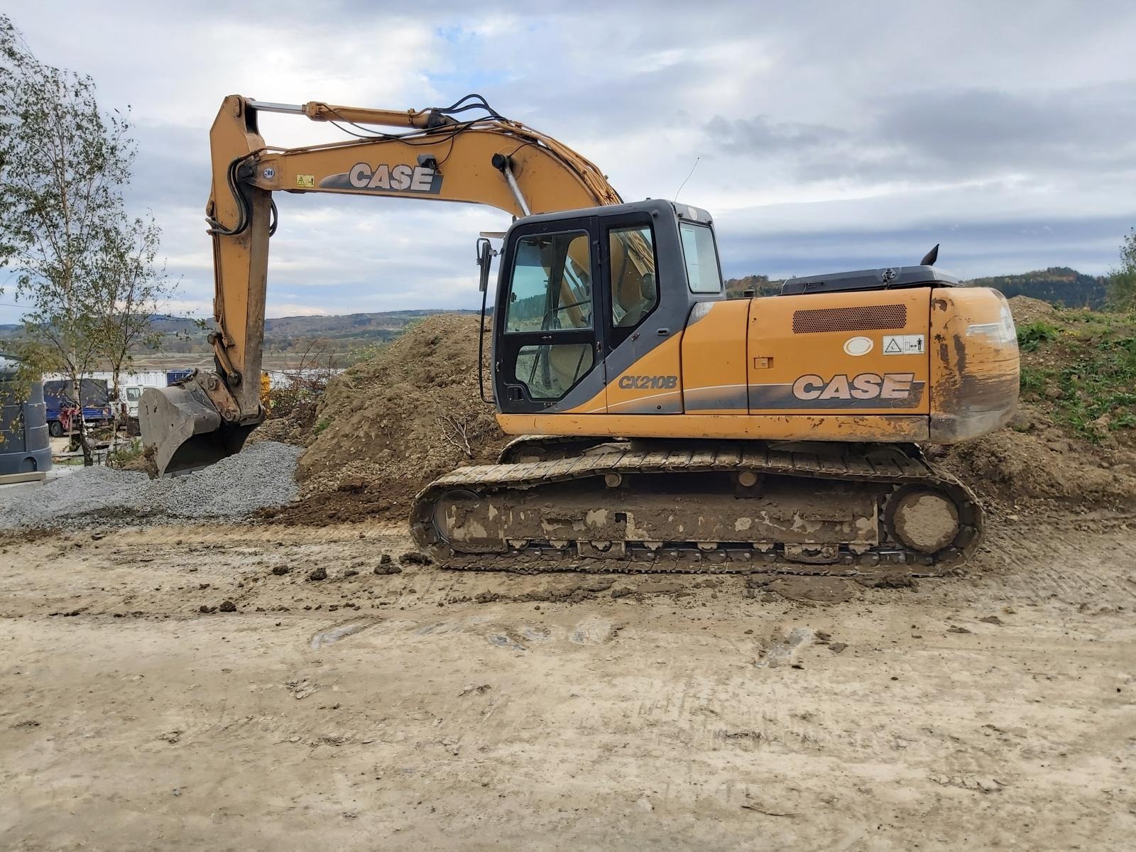 Pomarańczowa koparka CASE CX210B na placu budowy, z łyżką wypełnioną drobnym kruszywem, widoczne ślady błota na gąsienicach i dolnej części maszyny, w tle hałda ziemi i zalesione wzgórza...