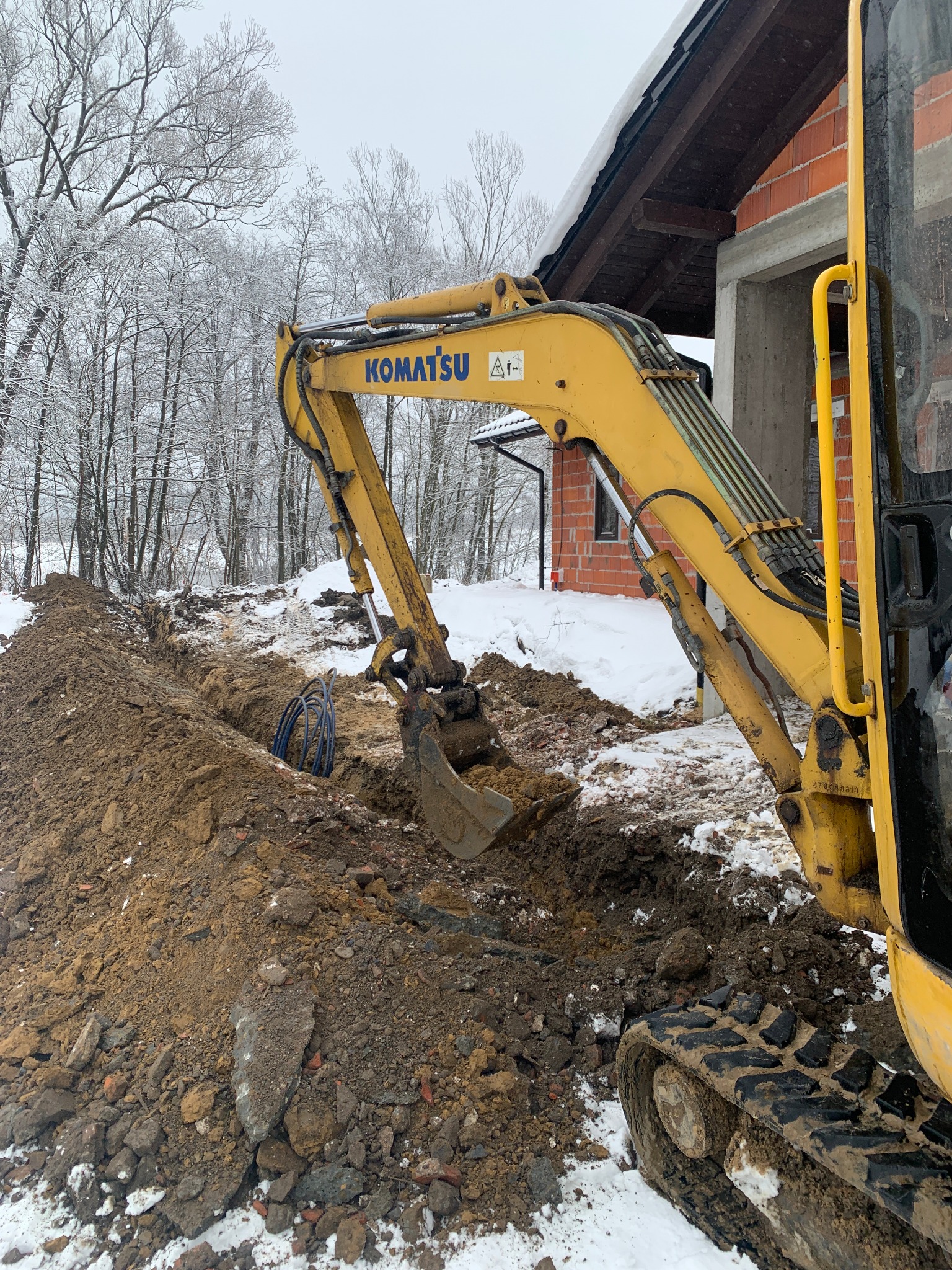 Żółta koparka Komatsu w trakcie wykonywania wykopu w ziemi, na tle ośnieżonych drzew i budynku w stanie surowym otwartym.
