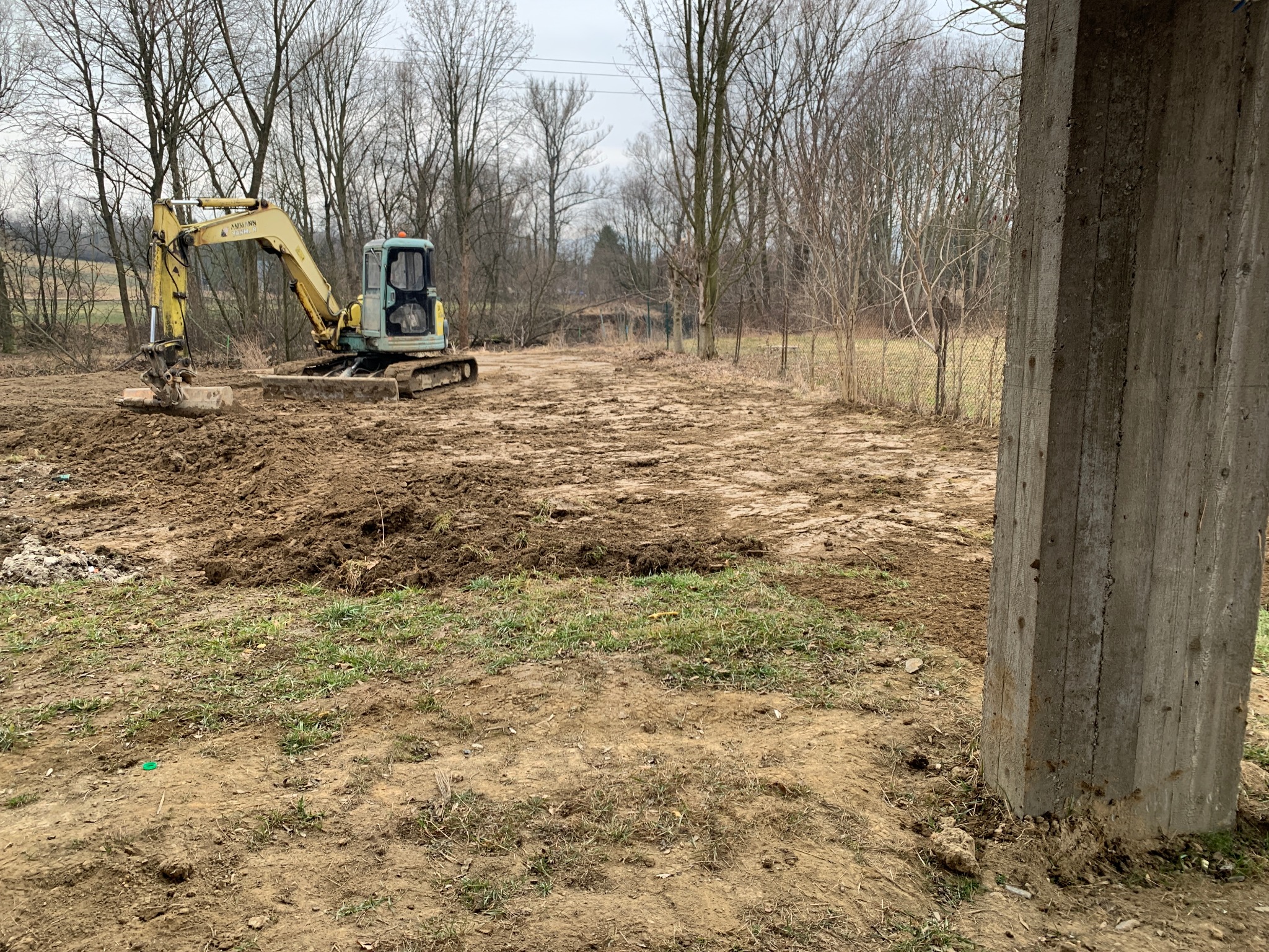 Żółta koparka na gąsienicach wyrównuje teren pod budowę, w tle drzewa bez liści i słup betonowy w prawym dolnym rogu.