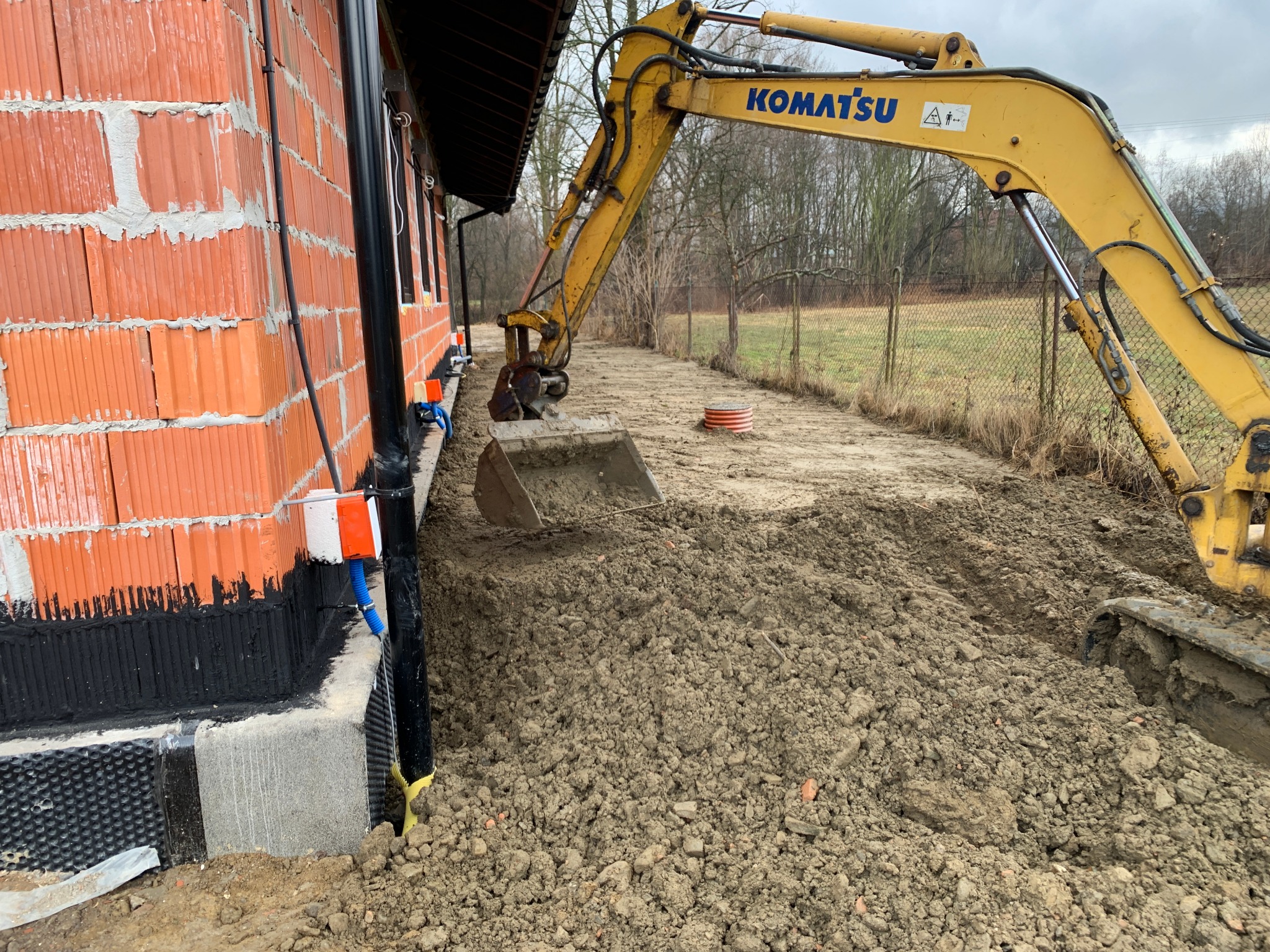 Żółta koparka Komatsu wykonuje prace ziemne przy nowo powstającym budynku z czerwonej cegły, widoczne przyłącza instalacji, teren wyrównany, w tle drzewa.