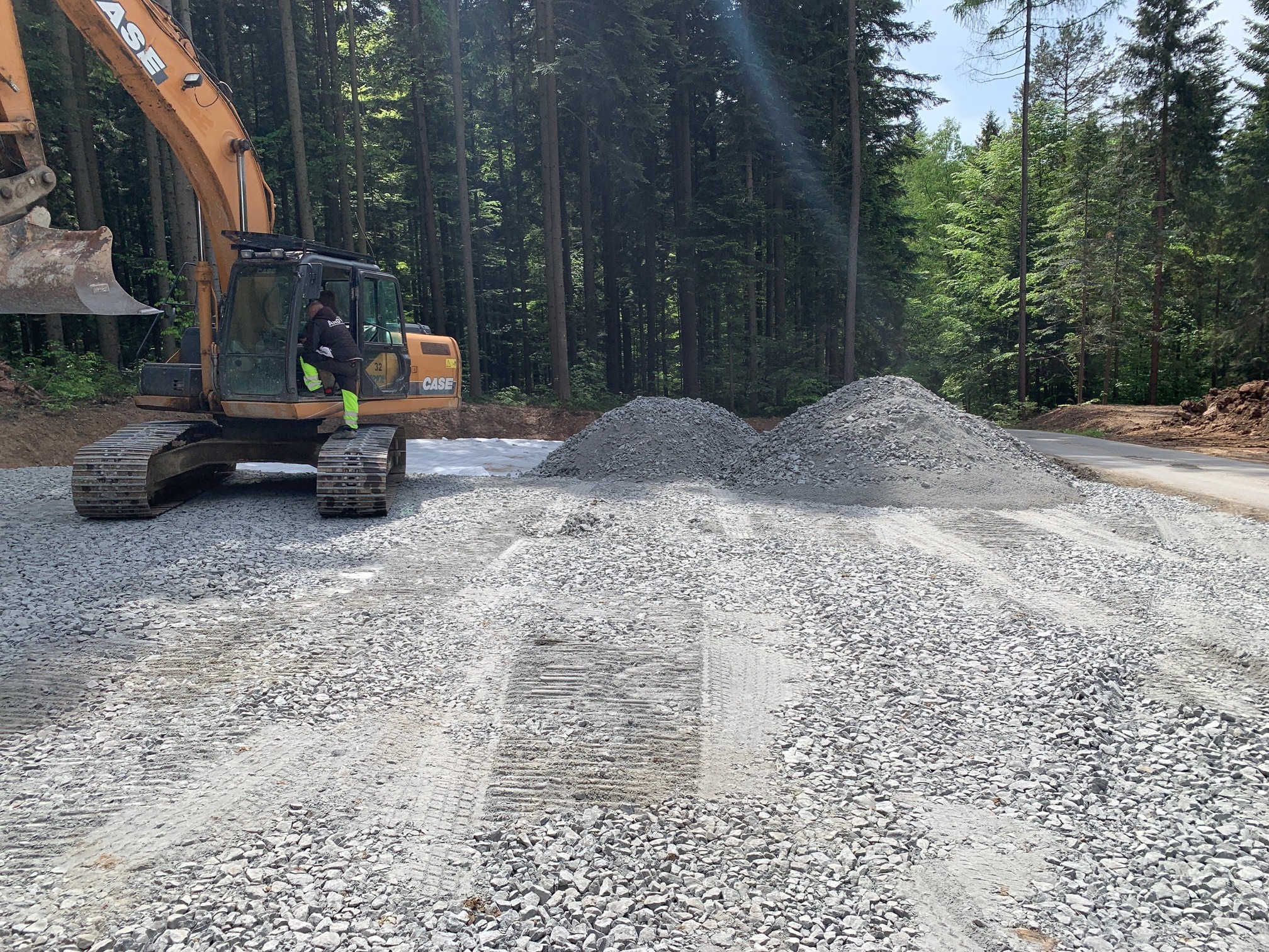 Pomarańczowa koparka CASE na placu budowy, przygotowująca teren pod drogę, widoczne dwie pryzmy szarego kruszywa, w tle las.