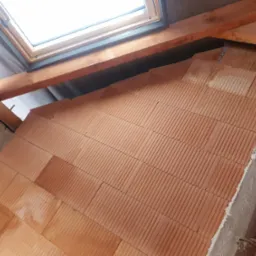 Wznoszenie ściany z czerwonej cegły ceramicznej pod oknem dachowym. Widoczne drewniane elementy konstrukcji dachu i fragmenty surowego betonu.