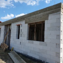 Kompleksowe usługi ogólnobudowlane - Ściana domu z betonu komórkowego w trakcie budowy, widoczne otwory okienne i wieniec. Na ścianie oparta drabina, w tle błękitne niebo.