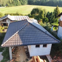 Kompleksowe usługi ogólnobudowlane - Nowy, grafitowy dach z dachówki ceramicznej na budynku w stanie surowym otwartym, z widokiem na wiejski krajobraz z zielonymi polami i drzewami.