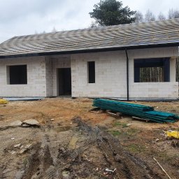 Kompleksowe usługi ogólnobudowlane - Surowy stan domu z białych bloczków, z widocznym dachem w trakcie budowy. Na pierwszym planie błoto i składowane deski, w tle las.