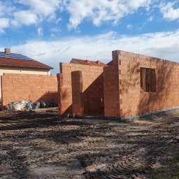 Kompleksowe usługi ogólnobudowlane - Wznoszenie ścian parterowego domu z bloczków ceramicznych. Widoczne otwory okienne i drzwiowe. Na dachu budynku obok panele słoneczne.