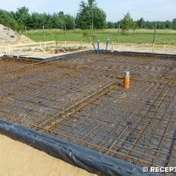 Kompleksowe usługi ogólnobudowlane - Zbrojenie fundamentów na czarnej folii, przygotowane do zalania betonem. Widoczne wystające rury instalacyjne i drewniane szalunki w tle. Dzień, teren budowy.