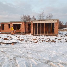 Kompleksowe usługi ogólnobudowlane - Budowa domu w stanie surowym zimą. Widoczne ściany z czerwonej cegły, drewniane podpory, śnieg i drzewa w tle. Częściowo ukończona konstrukcja.