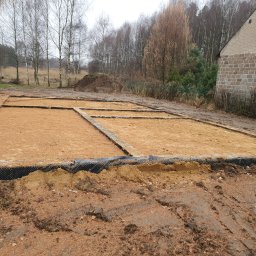 Kompleksowe usługi ogólnobudowlane - Przygotowany teren pod fundament domu z widoczną izolacją pionową i poziomą. W tle drzewa i fragment budynku z cegły.