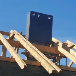 Niski kąt: surowa konstrukcja dachu z drewnianych belek i komin z otworami wentylacyjnymi na tle błękitnego nieba w słoneczny dzień. Etap budowy domu.