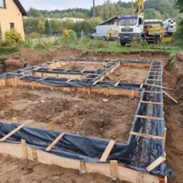 Szczegółowe ujęcie szalunków fundamentowych z widocznym zbrojeniem, przygotowanych do zalania betonem. W tle dom jednorodzinny i ciężki sprzęt budowlany.