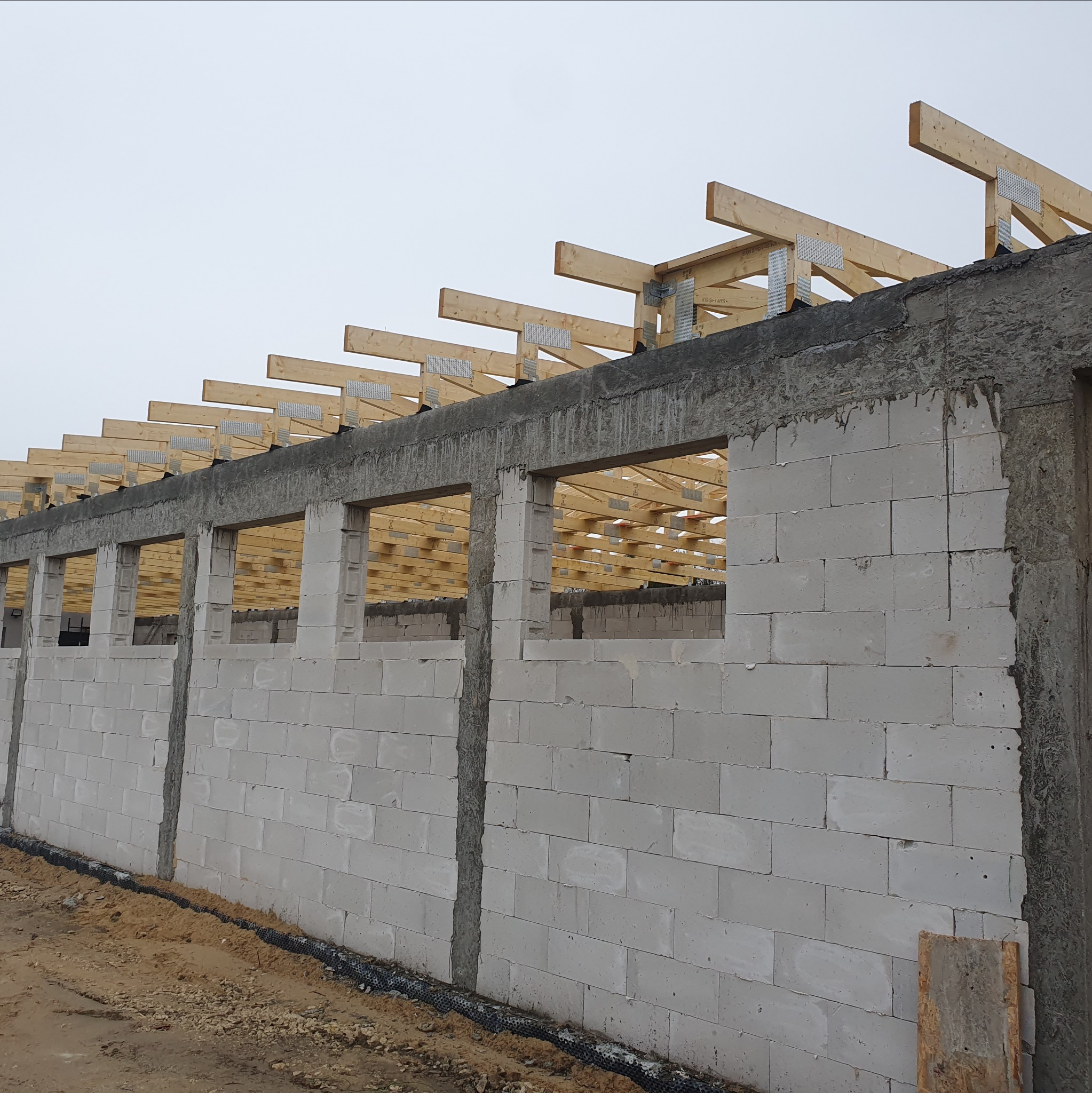 Budowa budynku z bloczków betonowych, widoczne ściany z otworami okiennymi i drewniana konstrukcja dachu. Etap wznoszenia ścian i montażu więźby dachowej.