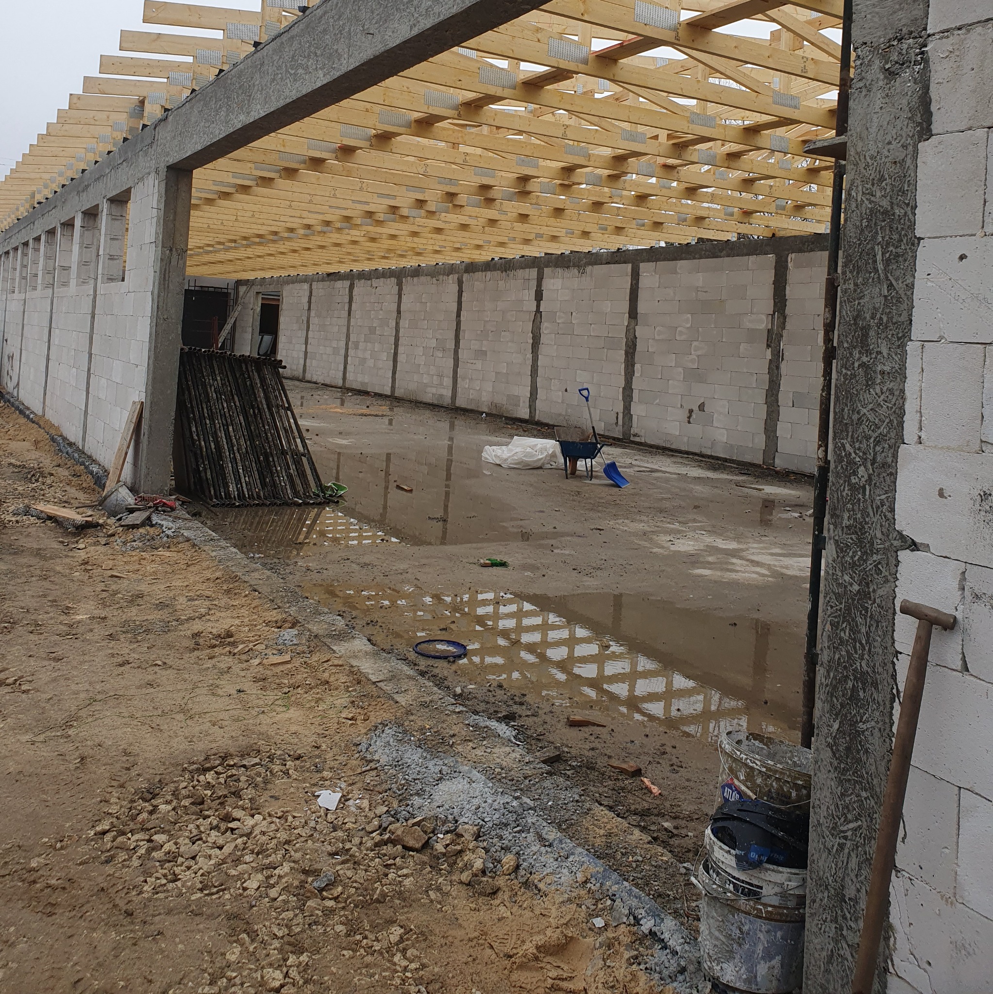 Budowa hali: ściany z bloczków, drewniana konstrukcja dachu, teren budowy z kałużami i narzędziami. Widok na wnętrze budynku w trakcie wznoszenia.