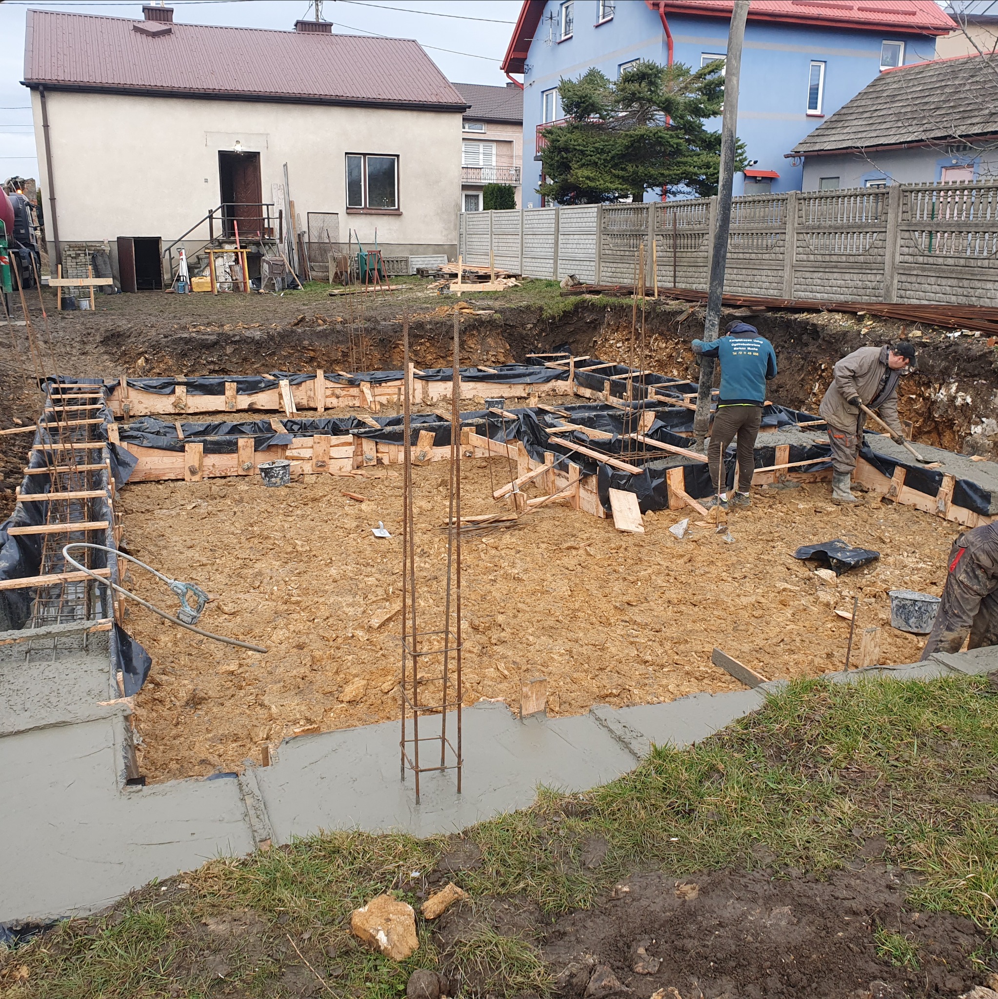 Wylewanie fundamentów pod dom: pracownicy rozprowadzają beton w szalunkach zbrojonych stalą. Widoczne budynki w tle i sprzęt budowlany.