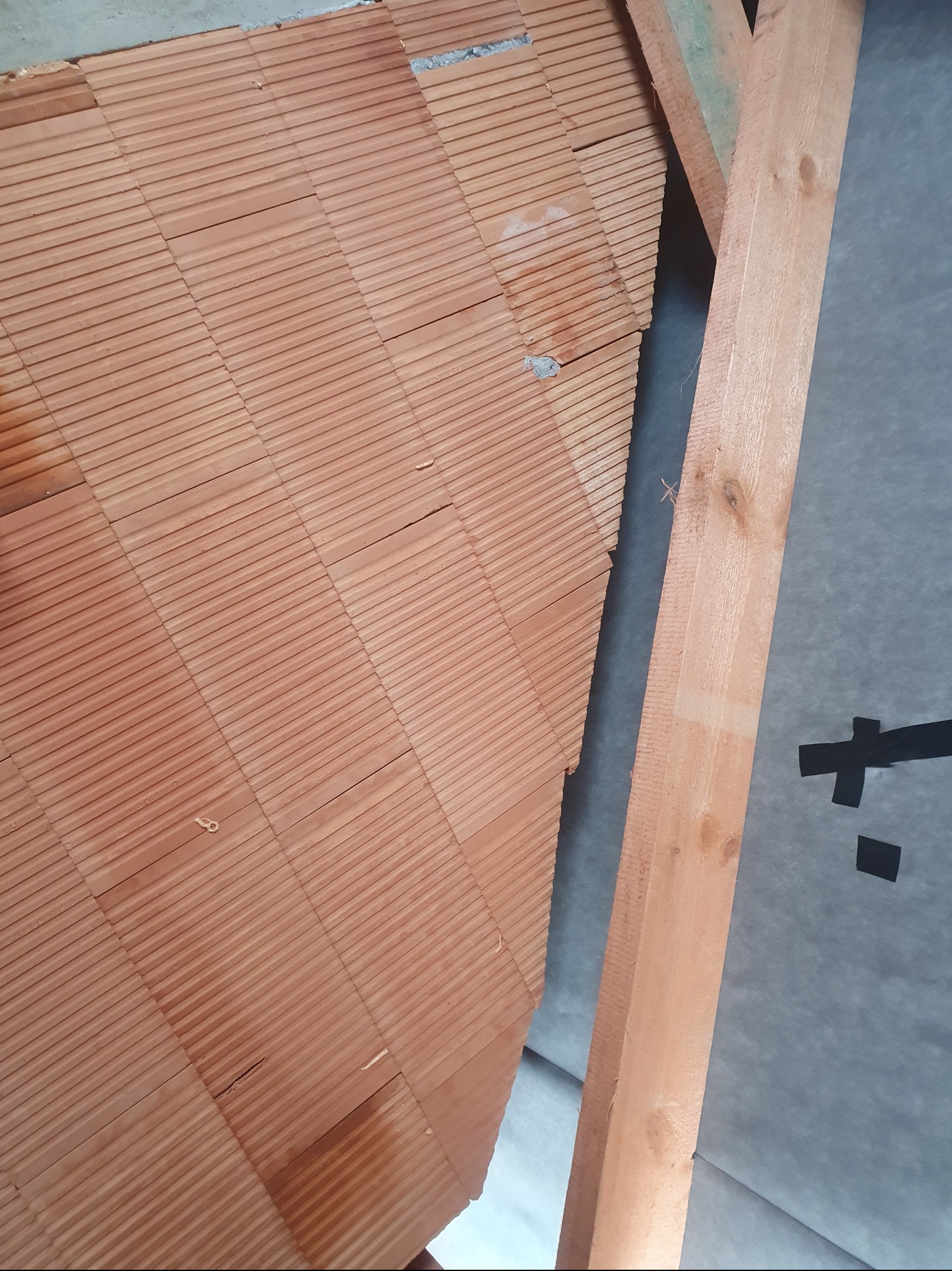 Ceglana ściana z pustaków ceramicznych, fragment drewnianej konstrukcji dachu i folia paroizolacyjna w tle. Widok z dołu.