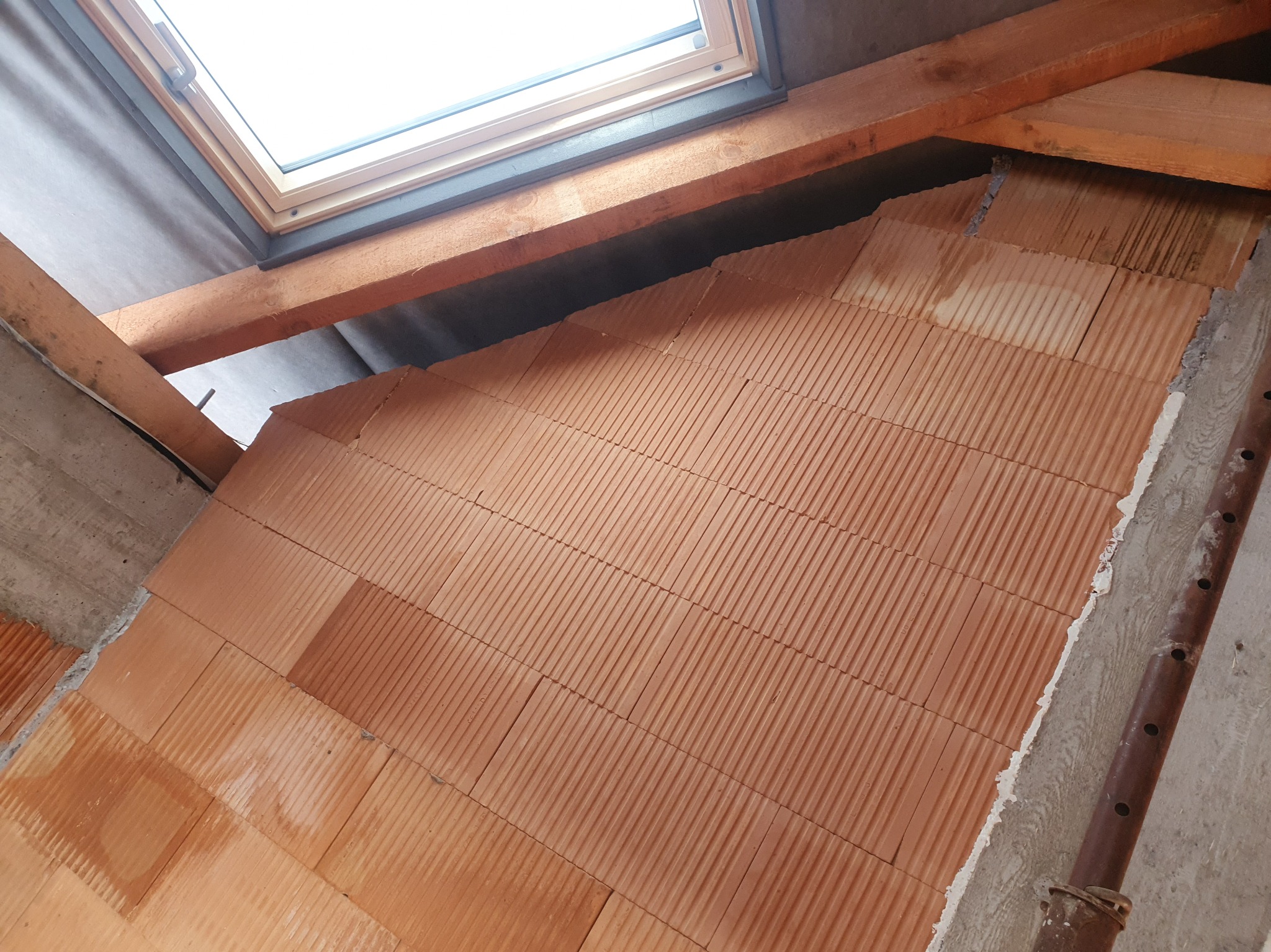 Wznoszenie ściany z czerwonej cegły ceramicznej pod oknem dachowym. Widoczne drewniane elementy konstrukcji dachu i fragmenty surowego betonu.