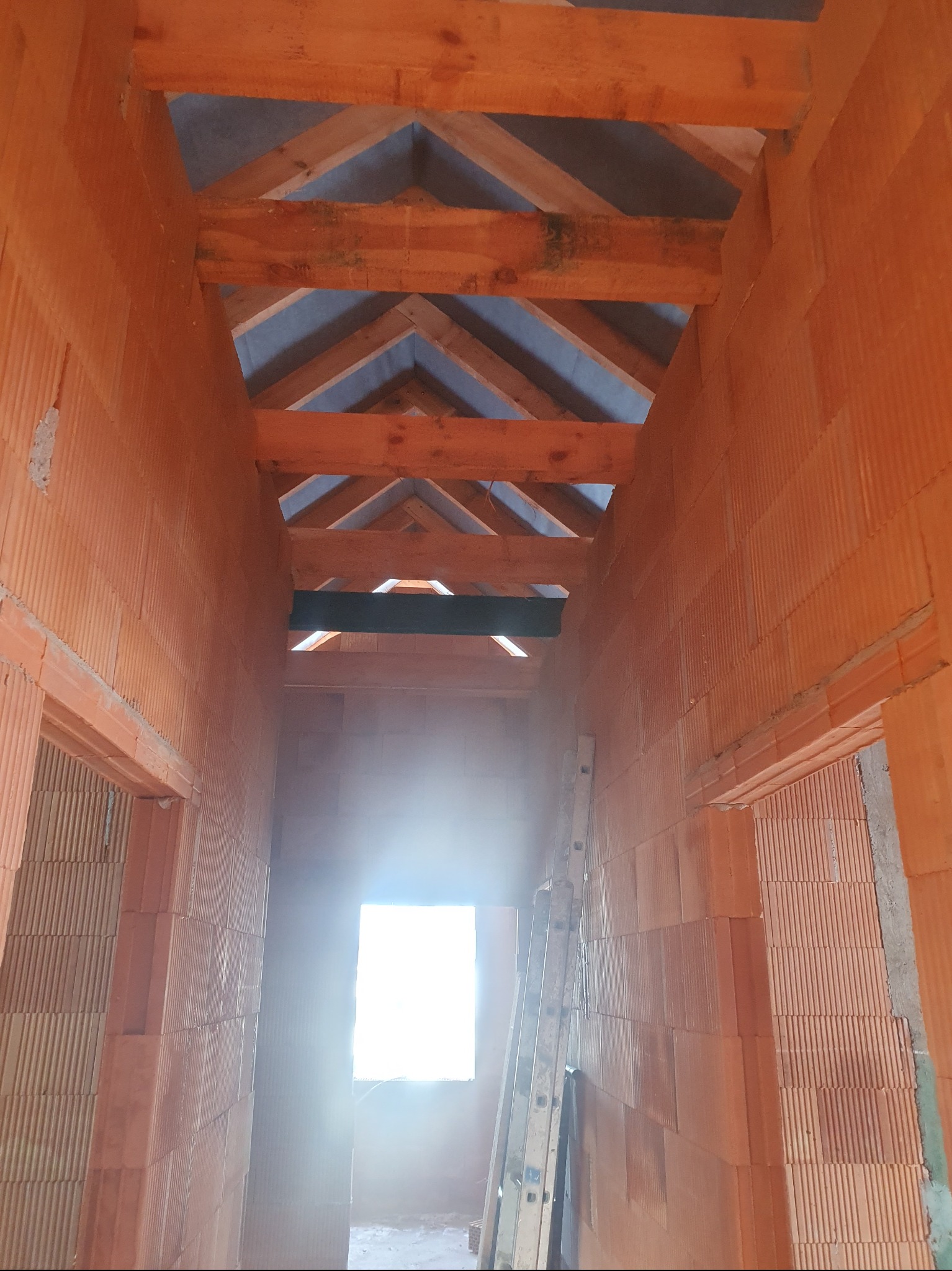 Wnętrze domu w trakcie budowy: ściany z czerwonej cegły, drewniany szkielet dachu i drabina oparta o ścianę. Widok na okno z intensywnym światłem.