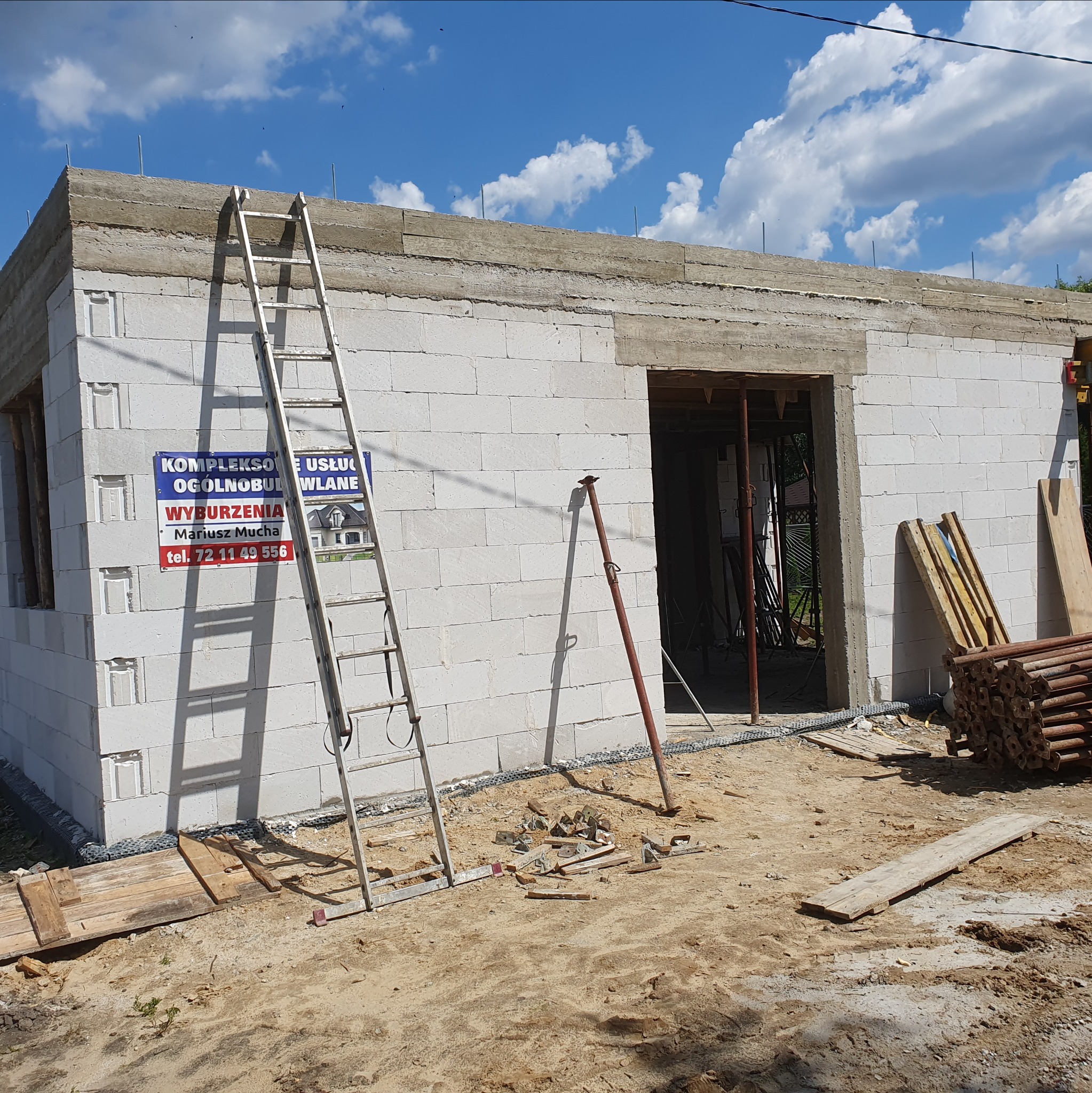 Wznoszenie ścian z białych bloczków betonu komórkowego, widoczna drabina oparta o ścianę, częściowo wylany strop oraz tablica informacyjna na ścianie. Etap budowy domu.